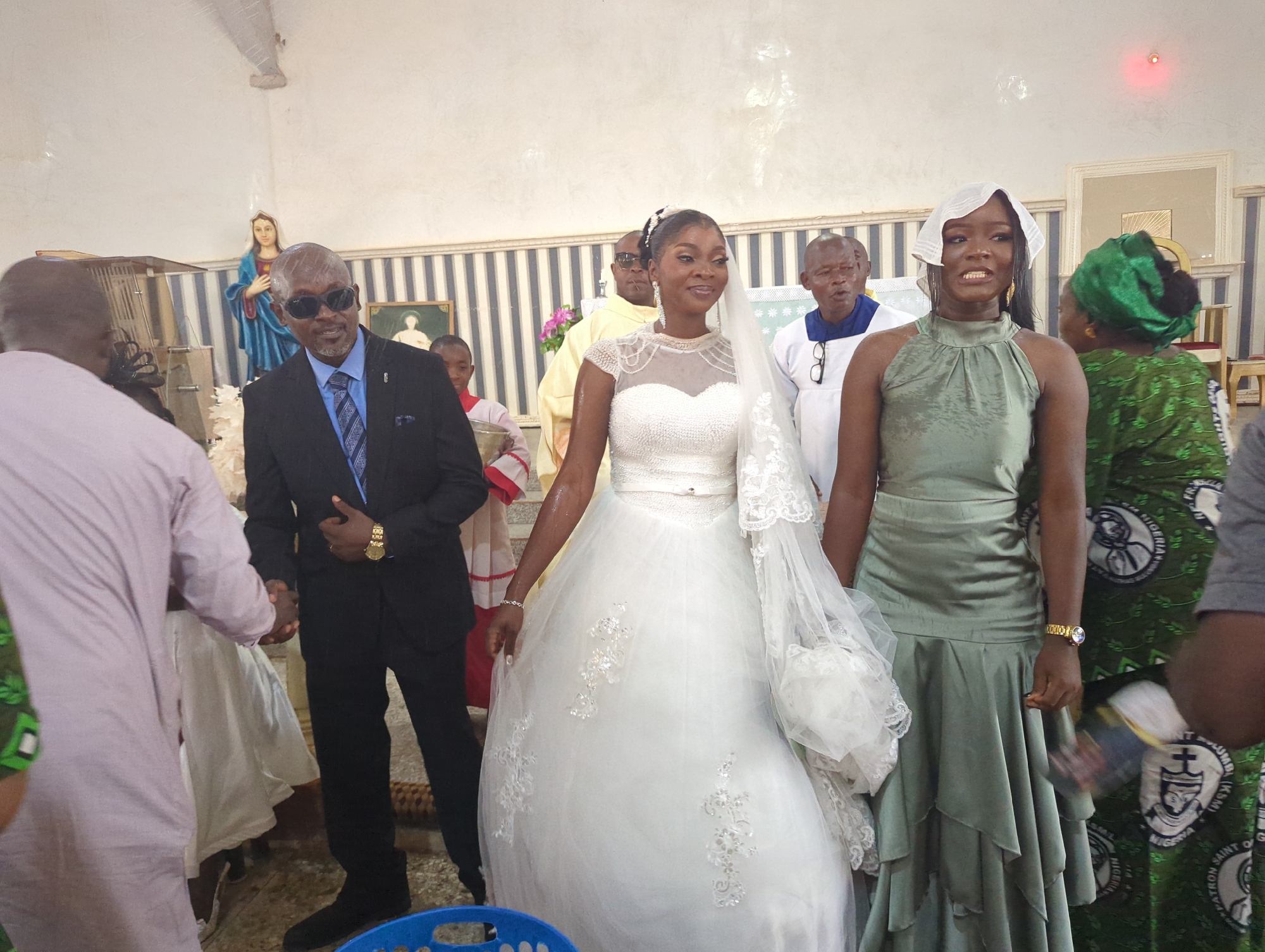 A wedding ceremony is taking place indoors in what appears to be a church. The bride, in a white lace wedding dress and veil, stands center, smiling. To her right is a woman in a green dress and headscarf, and to her left is the groom in a black suit. Several other people, including a man in a yellow clerical robe and a man in a white robe with a blue collar, are also present. The background features a statue of the Virgin Mary and a striped wall. The floor is tiled.