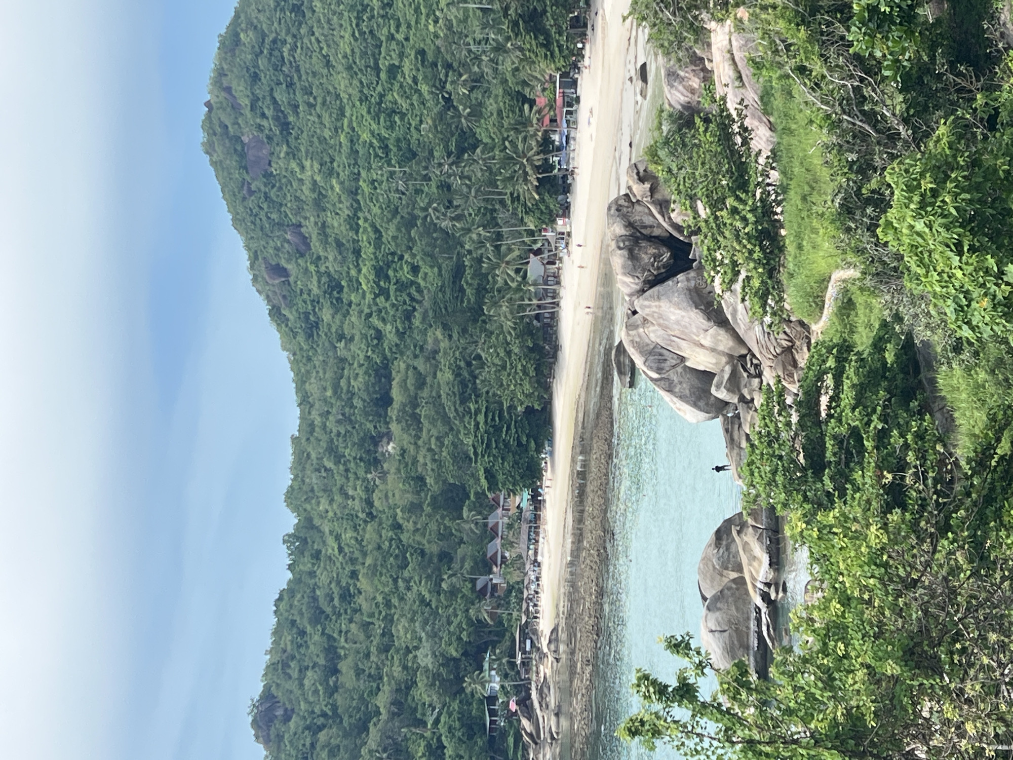 A medium shot depicts a scenic tropical bay with lush green hills rising in the background. The foreground features a rocky coastline with large, weathered boulders and dense green foliage. A person stands on a rock in the shallow, turquoise water. In the midground, a sandy beach is lined with palm trees and a few buildings, including some with red roofs. The water is calm and clear, with a few small waves gently lapping the shore. The sky is a soft blue with scattered clouds. The scene suggests a tranquil beach resort or a secluded bay in Thailand, possibly in the vicinity of Baan Chaweng Noi.