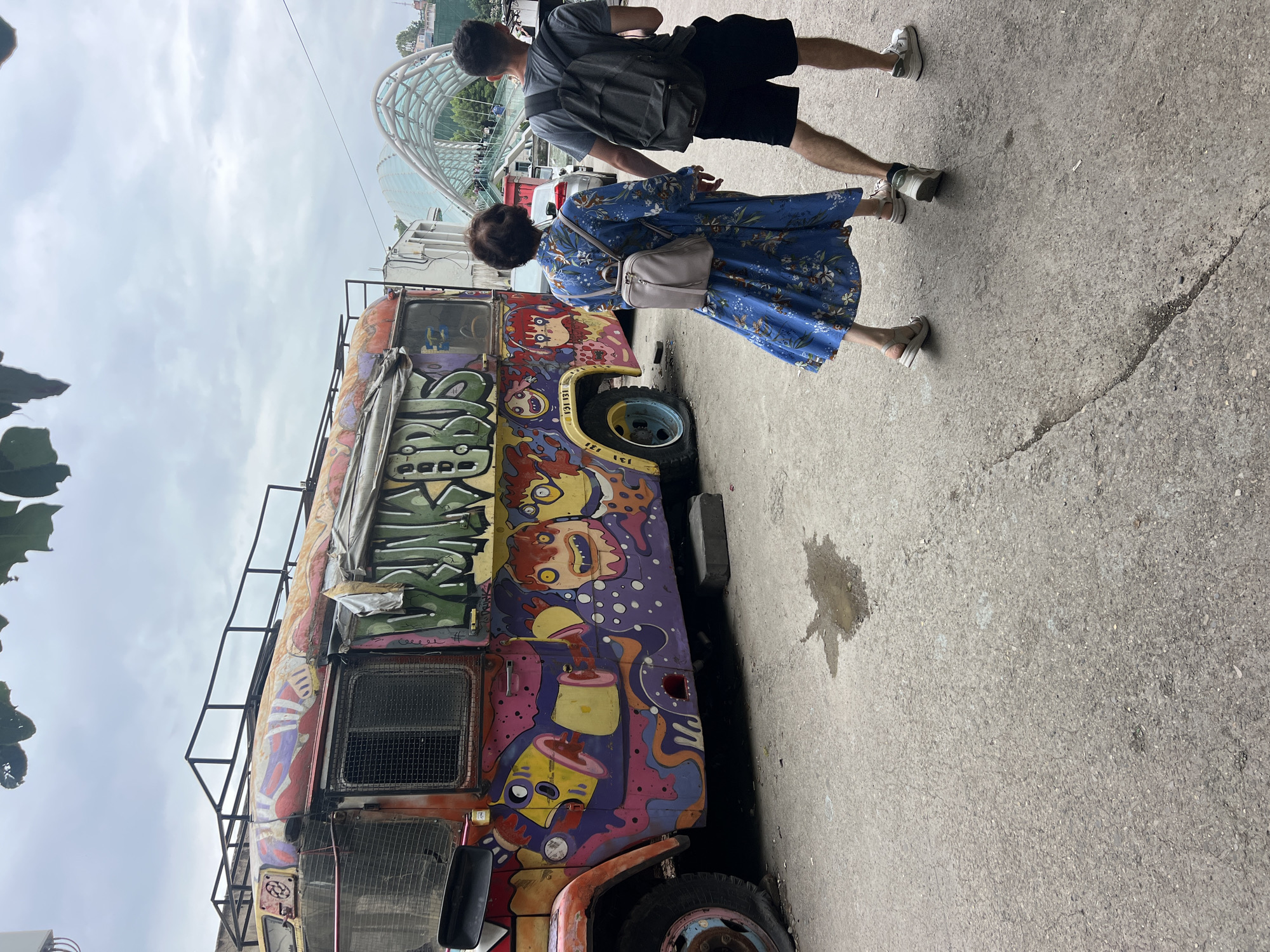 A man and a woman are walking away from the camera on a paved street in Tbilisi, Georgia, next to a brightly painted, graffiti-covered bus. The bus is old and appears to be in disrepair, with a flat tire on the front left side. The graffiti on the bus is colorful and abstract, with cartoon-like figures. The woman is wearing a blue floral dress and carrying a light gray backpack. The man is wearing a gray t-shirt, black shorts, and a dark gray backpack. They are holding hands. In the background, a modern pedestrian bridge with a distinctive arched design spans across a roadway. The sky is overcast.