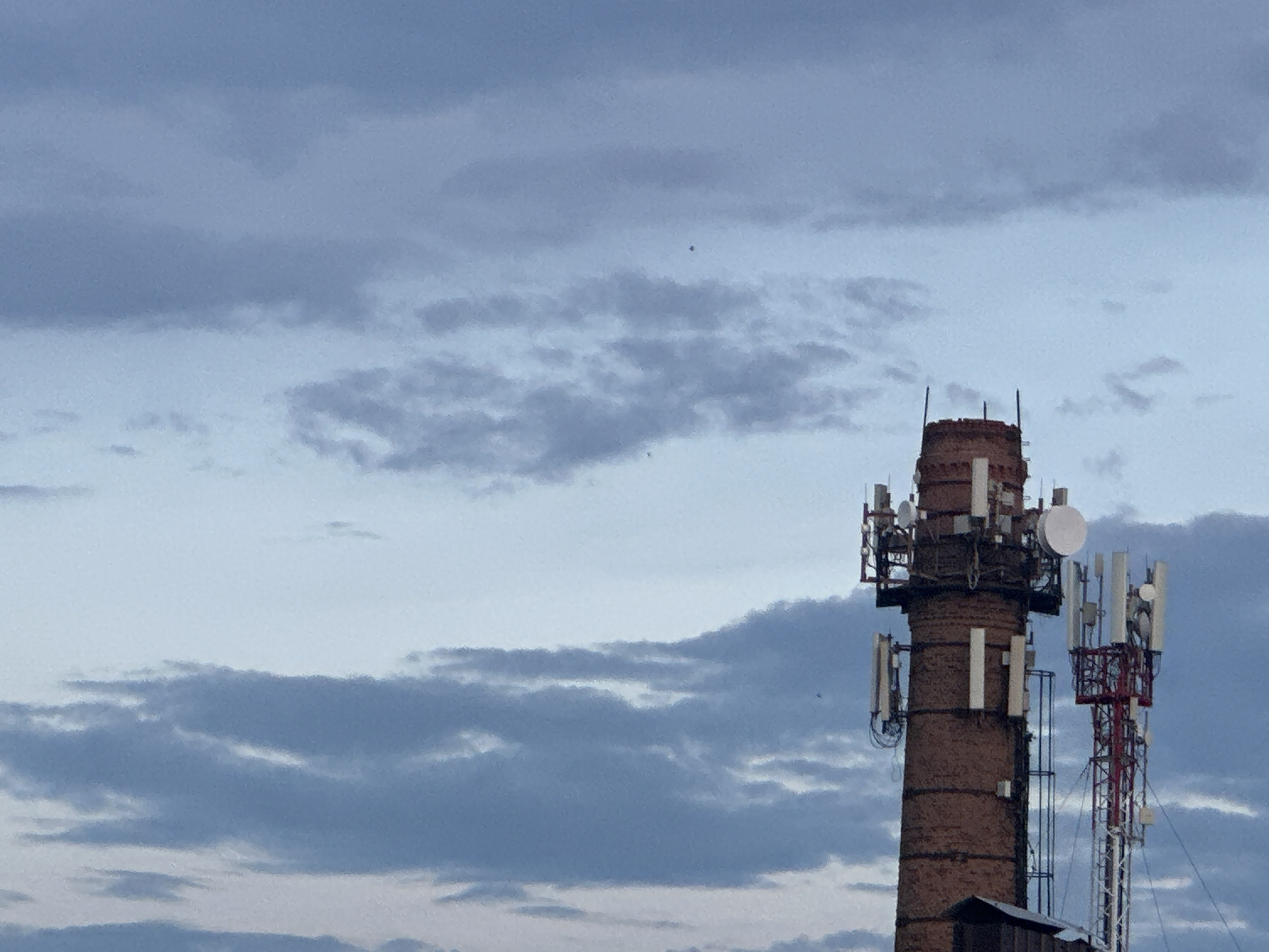 The image shows two cell towers against a cloudy sky. The tower on the left is a brick structure with multiple antennas and dishes attached. The tower on the right is a red and white lattice tower, also equipped with various antennas. The sky is filled with a mixture of gray and light blue clouds, suggesting it might be overcast or nearing dusk. The lower portion of the image shows a hint of a dark structure at the base of the brick tower, possibly a building or shelter. There are no people visible in the image. The scene appears to be an industrial or urban area where communication infrastructure is located.