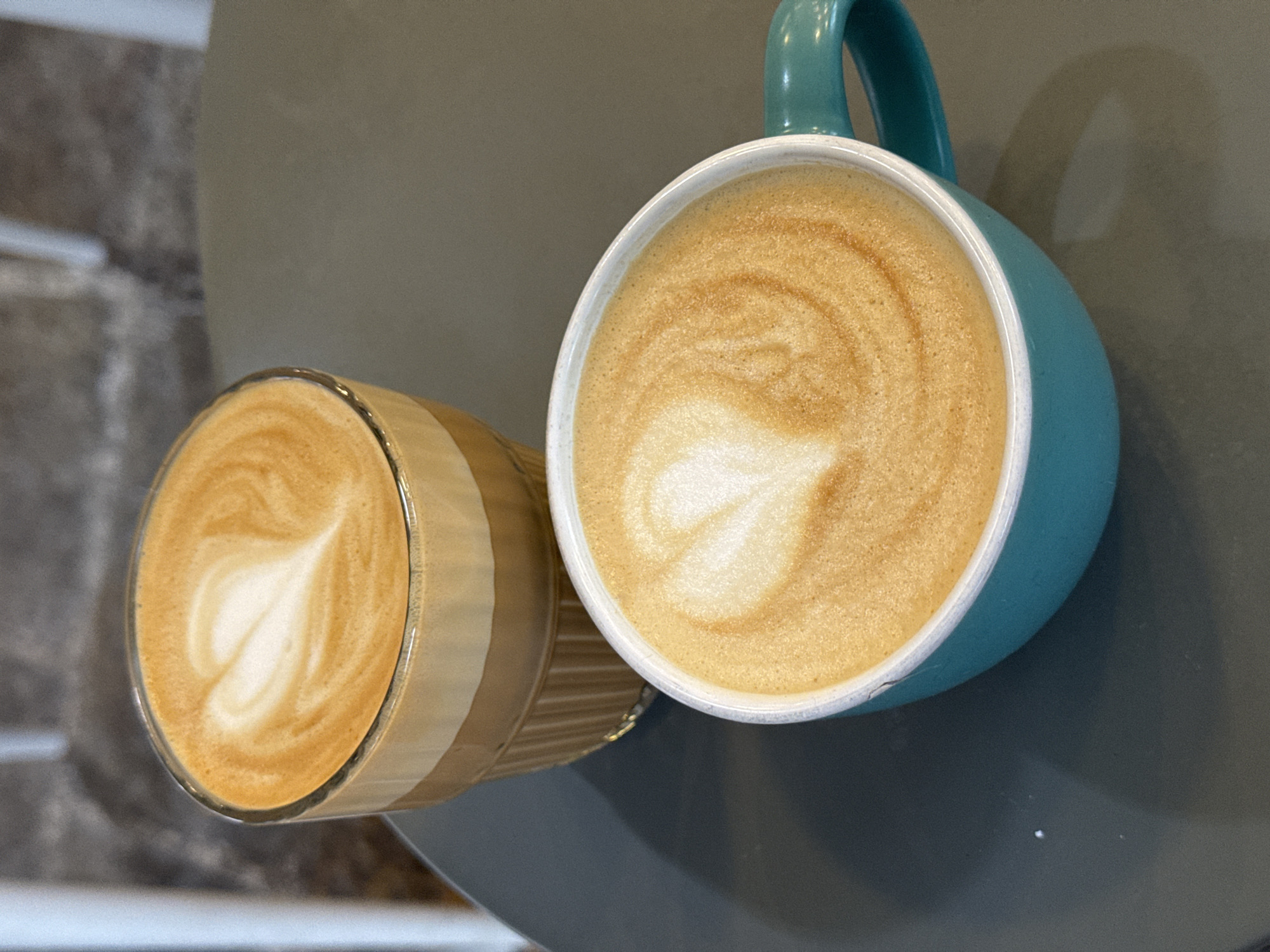The image displays an overhead shot of two coffee drinks, likely lattes, each featuring detailed heart-shaped latte art on their foamy surfaces. One drink is contained within a vibrant turquoise or teal ceramic mug with a visible handle and a crisp white interior rim. The other is presented in a clear glass cup, which allows the viewer to observe the distinct layers of coffee and milk below the textured foam.
Both cups are placed on a smooth, dark grey surface, presumably a table. The lighting is soft and even, emphasizing the rich, creamy texture of the foam and the warm brown tones of the coffee. The background is softly blurred, showing lighter grey tones and a subtle textured pattern on the left side, possibly a wall or another surface. No people are visible, and there is no discernible text within the image. The scene conveys a peaceful moment, likely in an indoor setting such as a cafe, with the focus solely on the attractively prepared beverages.