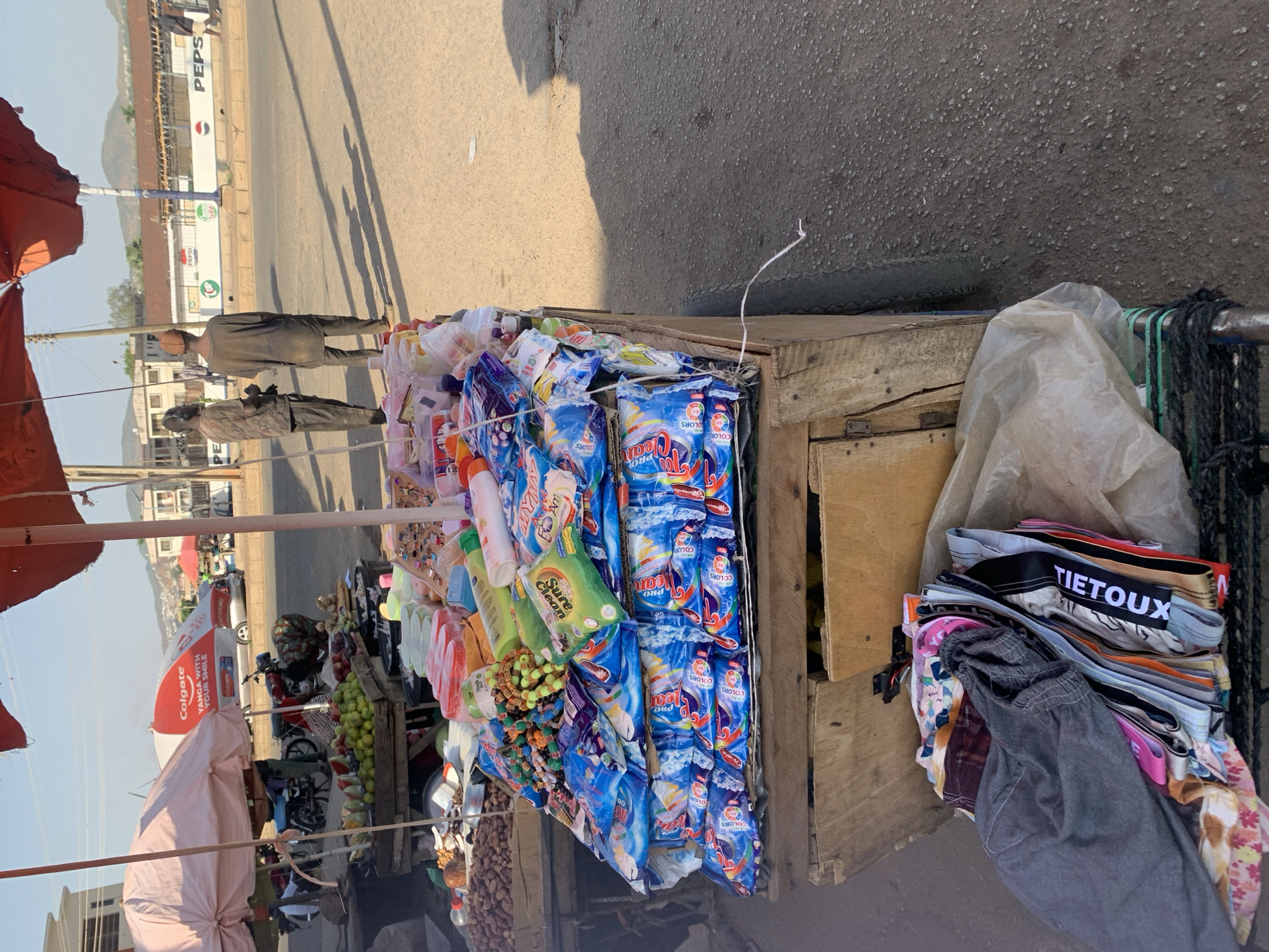 Here is a detailed description of the image:
**Setting:** The image is taken outdoors on a sunny day, possibly in a commercial or market area of New Stadium, Nigeria. A paved road or street is visible, along with buildings and trees in the background. The scene is adjacent to a roadside.
**Objects:**
* A wooden cart or stand is prominently featured. It is filled with various items, including toiletries (such as laundry detergent, toothpaste, and soap), as well as jewelry.
* The laundry detergent products are a prominent part of the display.
* A pile of folded clothing items is placed to the side of the cart, possibly for sale.
* Additional objects include a small basket of fruit, nuts and other items.
* A motorcycle is parked nearby.
**People:**
* Two people appear in the image.
**Environment:**
* The area appears to be a street or roadside in New Stadium, Nigeria.
* Several advertising umbrellas are visible.
**Overall Impression:** The image shows a roadside market stall. The stall is used to sell products like soap, clothing, jewelry and food.