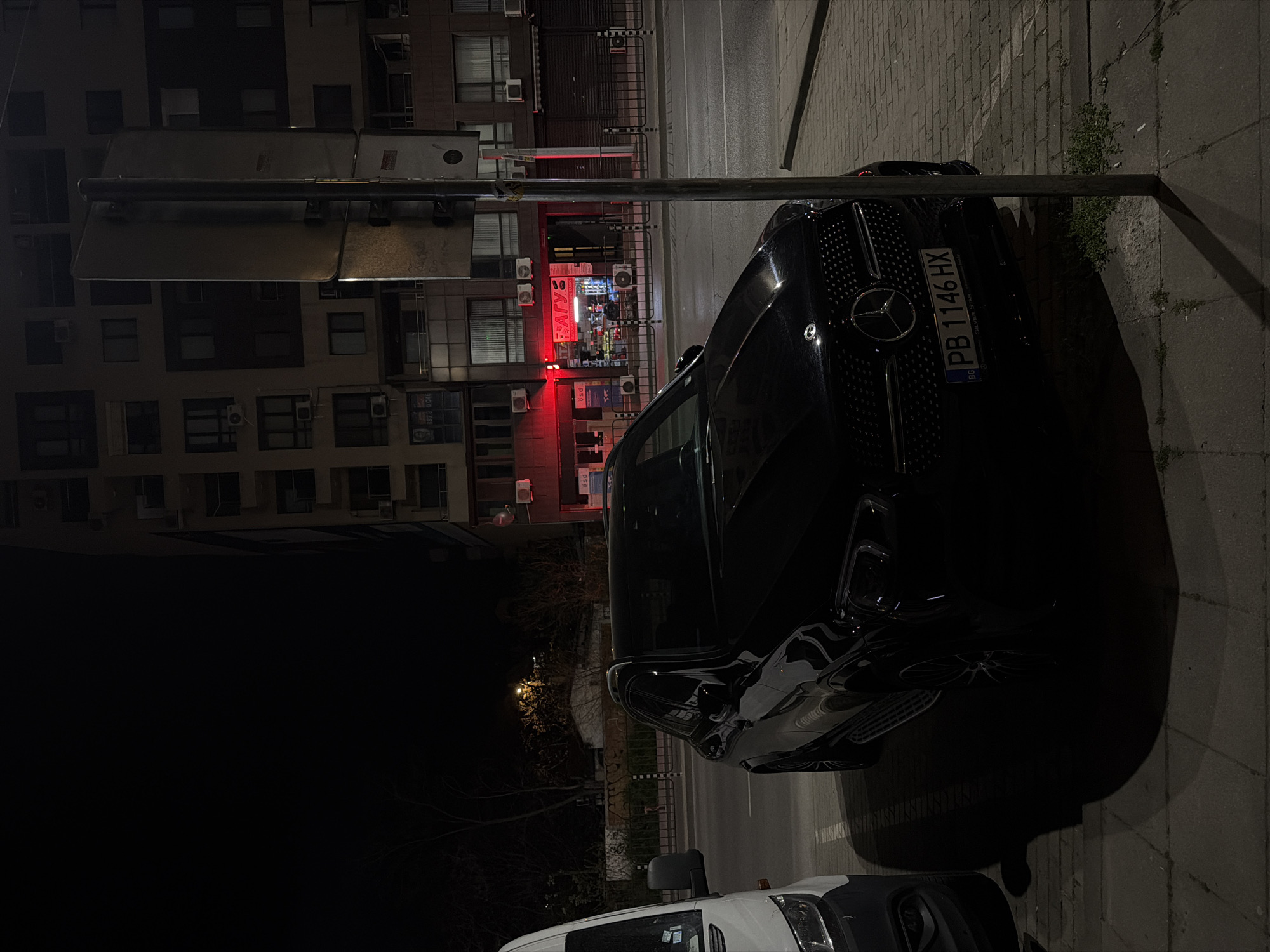 This nighttime image depicts an urban street scene in Plovdiv, Bulgaria. The primary subject is a black Mercedes-Benz car, parked on the right side of the frame, facing away from the viewer. Its rear license plate, "PB 1146 HX," with the "BG" country code and "Silver Star" on the frame, is clearly visible. The front portion of a white van is also partially visible in the immediate foreground, below the Mercedes.
A prominent street pole stands to the left of the Mercedes. Across the paved street and sidewalk, a large, multi-story building forms the backdrop. The ground floor of this building features a small, brightly lit shop with red and white illumination. The shop's sign displays Cyrillic text, including "АТГУ" (ATGU) in red. Other smaller red signs with partial text like "psa" are visible near the entrance. Above the shop, a section of the building appears to have a rolled-down metal security gate. The building's facade is characterized by numerous windows, some balconies, and several air conditioning units. A poster or sign on the building has the numbers "3873 204" printed on it.
The sidewalk consists of tiled sections, with small weeds growing in the cracks. The overall lighting is dim, with artificial lights from the shop and street lamps providing sparse illumination, creating reflections on the cars' surfaces. No people are visible, contributing to a quiet, nocturnal urban atmosphere.