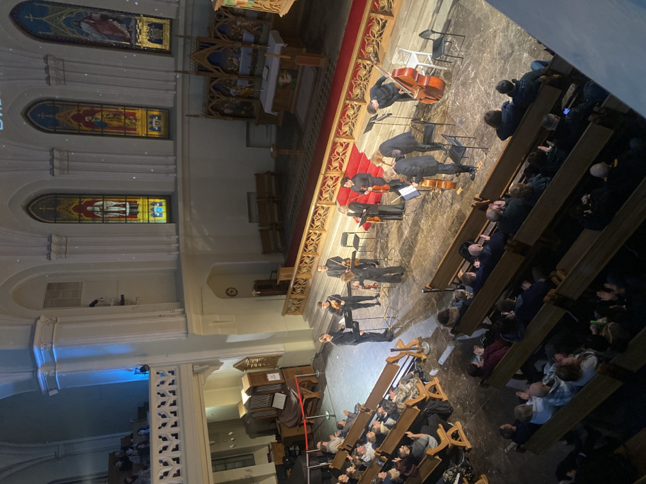 This image captures a classical music concert taking place inside the Evangelical Lutheran Cathedral of Saints Peter and Paul in Moscow, Russia.
The scene is viewed from an elevated balcony, looking down into the main nave. Nine musicians, dressed in dark formal attire, are positioned on the central floor area, performing with string instruments including violins, cellos, and a double bass. Music stands are placed in front of each musician. One male musician stands prominently in the center-left, facing the audience, possibly a conductor or soloist.
A substantial audience occupies numerous wooden pews on the main floor, facing the performers. Many individuals are visibly clapping, indicating appreciation for the performance. Additional spectators are present in a balcony section on the left.
The cathedral exhibits Gothic architectural features, including high ceilings, pointed arches, and sturdy pillars. Three large, vibrant stained-glass windows depicting religious figures are visible on the far wall above the altar. The altar area, located on a raised platform at the far right, features a white altar cloth, a small cross, and an intricately carved wooden reredos. A red carpet covers the elevated platform leading to the altar. A large, wooden pipe organ is situated on the left side of the main floor, partially obscured by the balcony. Blue accent lighting illuminates one of the pillars on the left. The general lighting suggests an evening event.