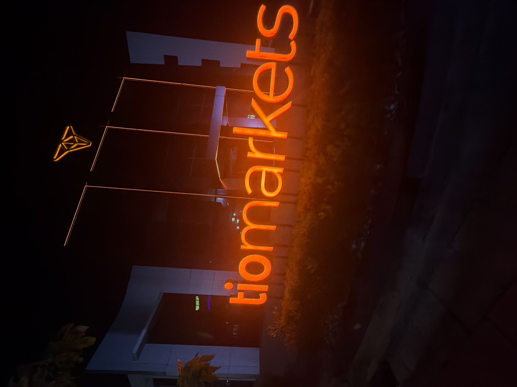 In the city of Agios Athanasios Municipality, Cyprus, an outdoor nighttime scene depicts a building facade with a large, bright orange neon sign spelling out "tiomarkets." The sign is illuminated and stands out against the dark surroundings. Above the sign, a smaller, stylized orange logo is visible. The building itself features white architectural elements and large glass windows that reflect the neon light. In front of the building, there is a dark sidewalk or road and some low-lying vegetation, also illuminated by the ambient orange glow. The overall impression is that of a commercial establishment or office building with prominent branding.
