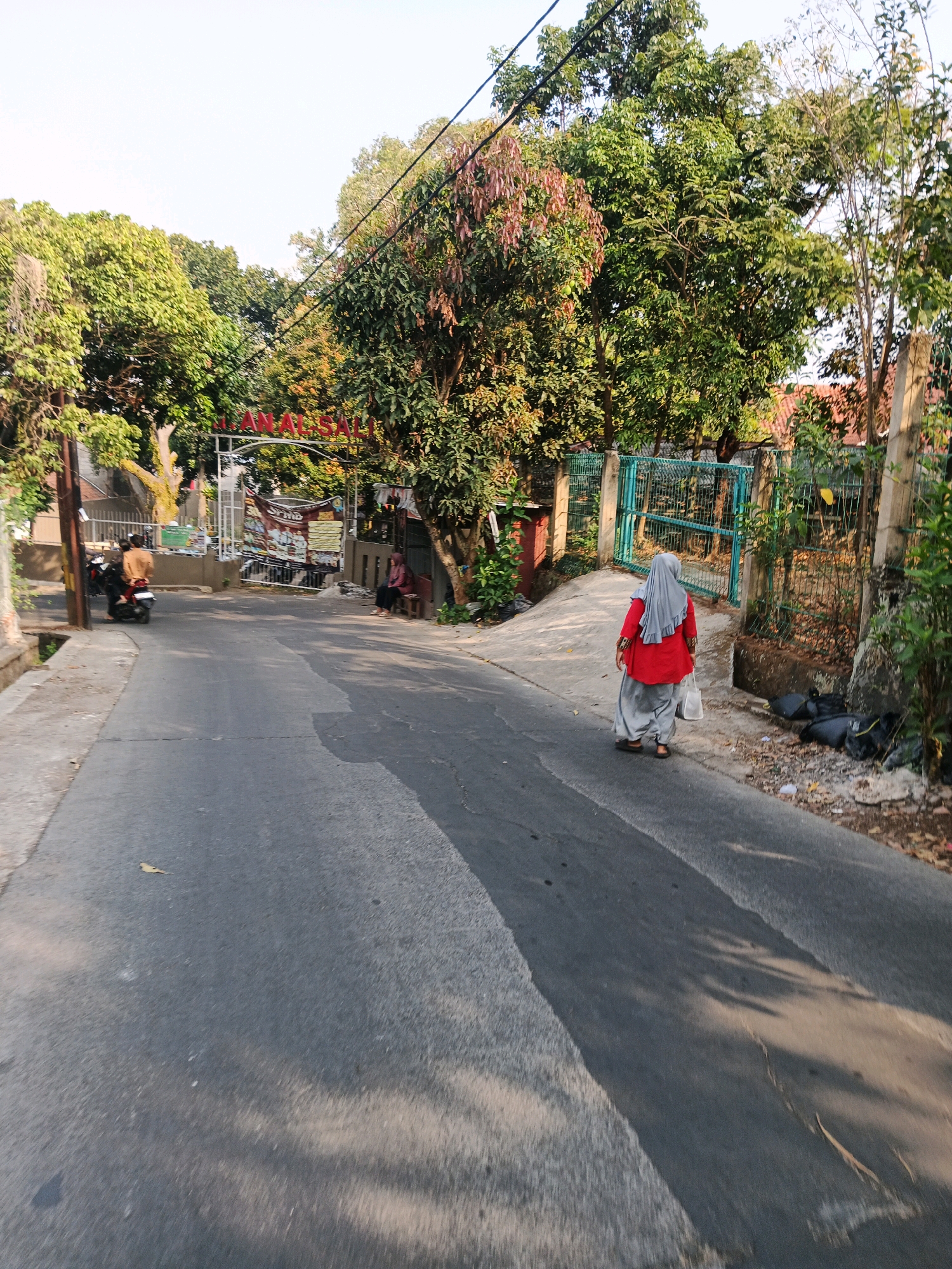 The image captures a street scene in Bandung City, Indonesia, on a bright day with sunlight casting shadows. In the foreground, a paved road runs diagonally from the bottom left towards the center. To the left of the road, a grey concrete retaining wall is visible, with a drain at its base. Further up the road on the left, a person on a motorcycle wearing a brown jacket is partially visible.
The right side of the road is bordered by a green metal fence. Behind the fence, a person wearing a red top and a grey headscarf walks away from the camera, carrying a white plastic bag. There are several black garbage bags piled up near the fence. Further back, a white archway with red lettering, possibly a street sign, is visible amidst lush green trees and foliage. Two people are seated on a bench to the right of the archway. The environment suggests a suburban or residential area with ample greenery. The overall impression is one of a typical day in a Indonesian city.