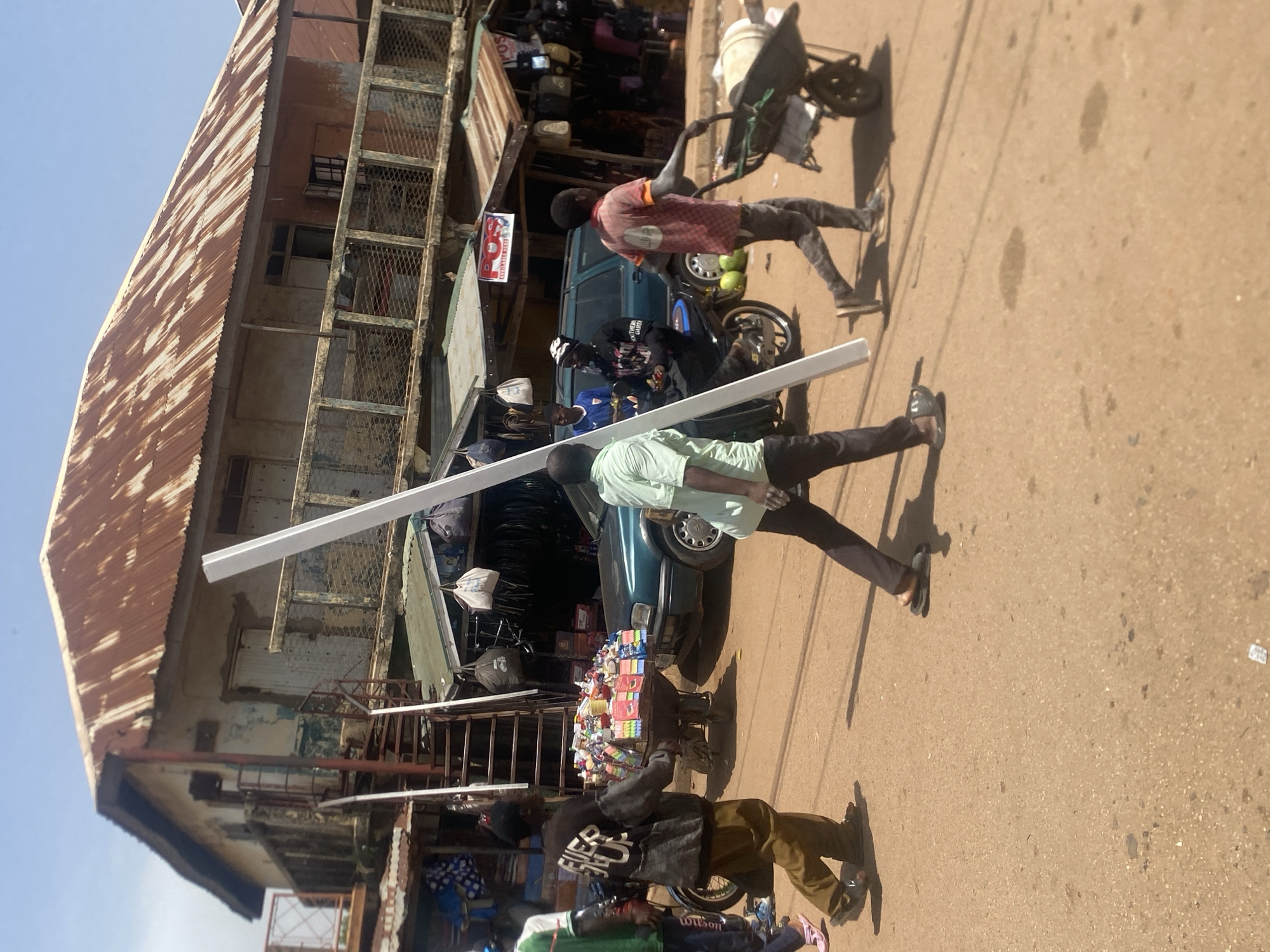 In Bukuru, Nigeria, on a dusty, sunlit street, a group of individuals are engaged in various activities. In the foreground, a man wearing a light green shirt and dark pants walks to the left, holding a long, white rectangular object. To his left, another man in a black jacket with "EVER UP" written on the back and brown pants walks to the left, pulling a cart laden with colorful candies and snacks. Behind them, a green SUV is parked on the street.
In the background, a dilapidated, two-story building with a rusty corrugated iron roof looms over the scene. A metal staircase leads up to a balcony with a railing. The building's facade is weathered and peeling, with visible signs of age and neglect. Several market stalls with awnings are set up along the street, displaying various goods. A man in a red and orange checkered shirt pushes a wheelbarrow loaded with cylindrical objects to the right. Another person, wearing a blue shirt and a white patterned headscarf, sits on a motorcycle. The overall impression is one of a bustling, informal marketplace in a developing urban environment.