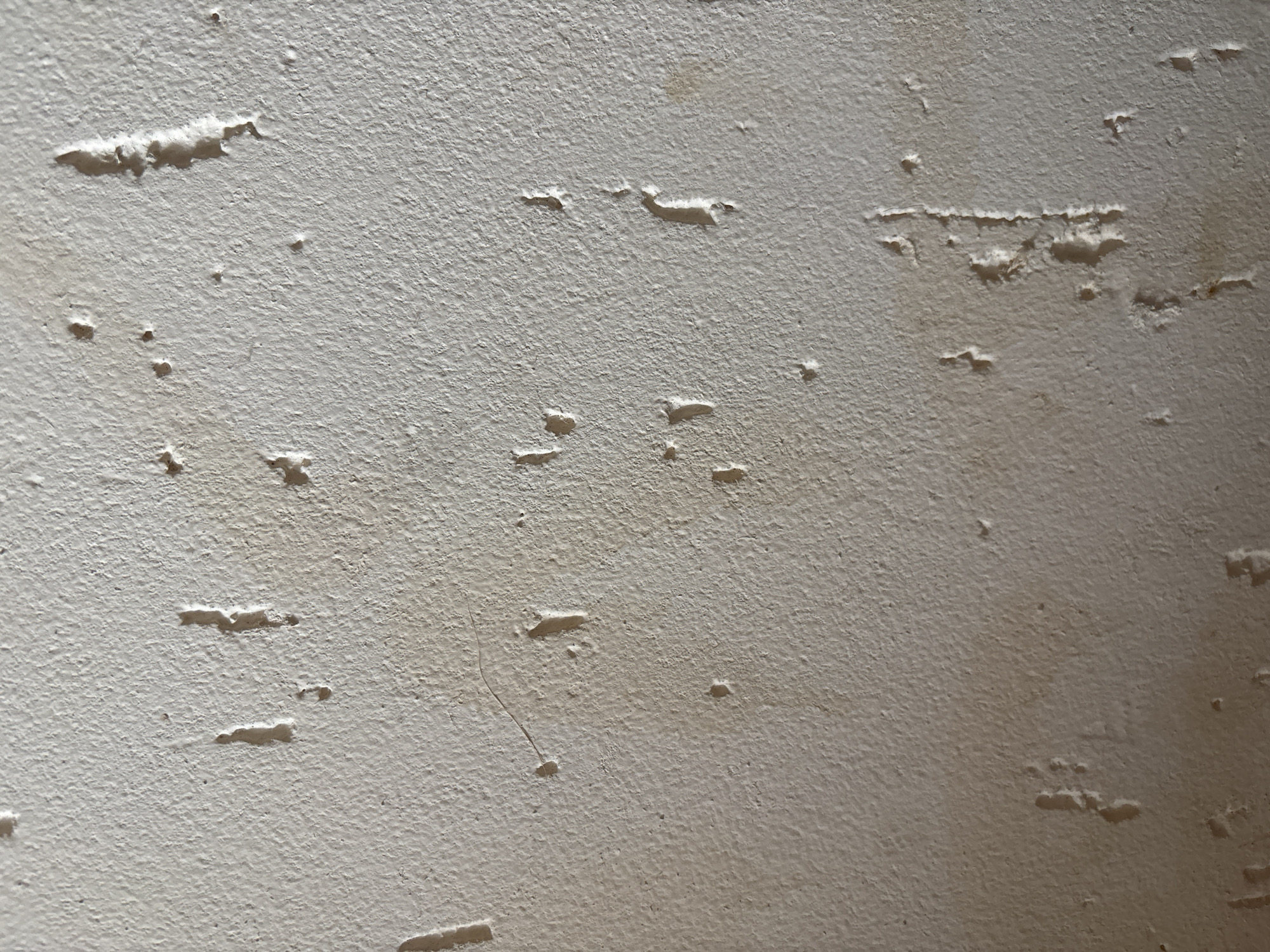 The image shows a close-up, dimly lit view of a textured, off-white wall surface, likely painted plaster or stucco. The surface is uneven, with numerous small pits, divots, and some larger, elongated indentations that appear to be imperfections or damage to the material. There are also a few faint, dark smudges or stains scattered across the wall, particularly in the lower portion of the frame. The lighting casts subtle shadows, highlighting the texture and irregularities of the wall. The location is described as Відамлянскі сельскі Савет, Belarus, suggesting this is an interior or exterior wall in a rural area. There are no people, animals, or other identifiable objects present in the frame.