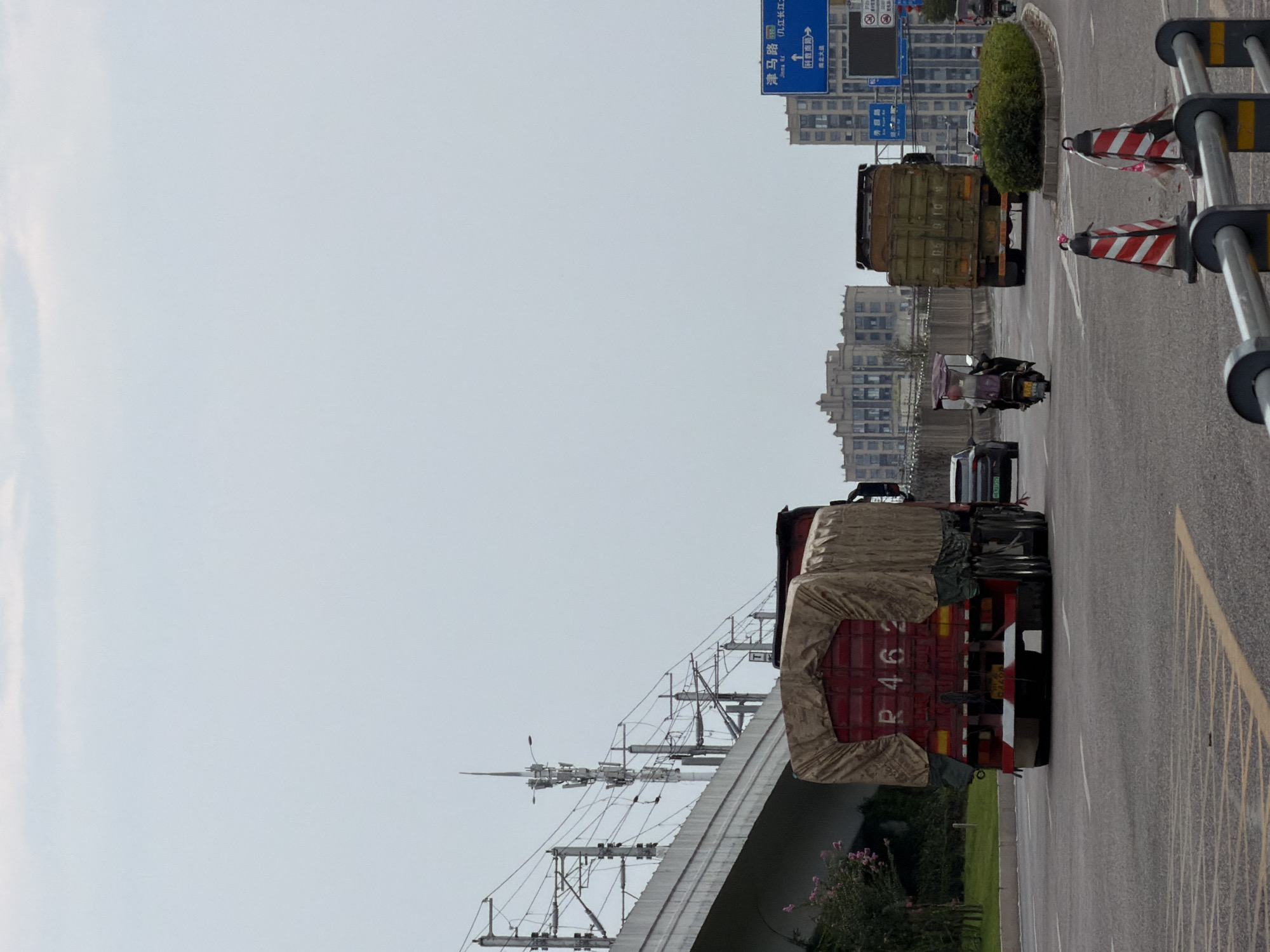A medium shot captures a street scene in the city of Jiangjin, China, under an overcast sky. In the foreground, a red truck with a covered cargo bed, displaying the license plate "R 462", is positioned on the right side of the road. Ahead of it, a motorcycle with a passenger navigates the traffic. Further down the road, a green truck is visible. The background features modern buildings and overhead power lines, suggesting an urban environment. Road signs with Chinese characters indicate directions to "Jingma Road" and other locations. The road is paved and marked with lane dividers. Several traffic cones are placed on the road, indicating potential construction or a road closure. The overall scene depicts everyday transportation and infrastructure within the city.