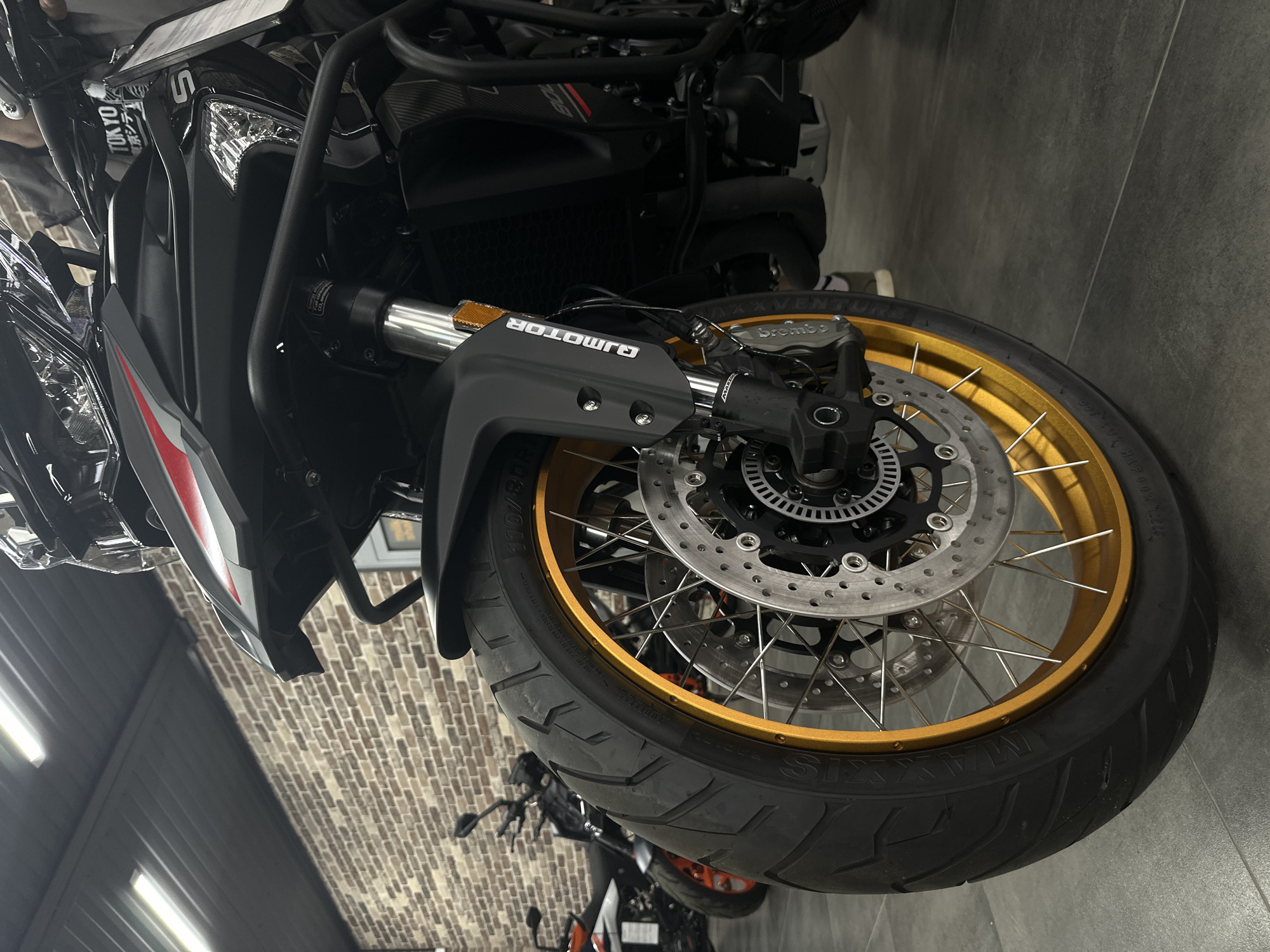 This image depicts the interior of a motorcycle showroom in Rostov-on-Don, Russia. The primary subject is a black QJMotor adventure-style motorcycle positioned prominently in the foreground, angled slightly to the left. Its large front wheel features a gold-colored spoked rim and a black tire with visible tread, marked with "110/80" and other textual details. The front suspension consists of silver stanchions and black lower fork guards, the latter bearing "QJMOTOR" branding and an orange reflector. Dual silver disc brakes are visible, with a black Brembo caliper on the left. The motorcycle's front fairing is black with red and white striped accents and a clear windscreen. Black protective crash bars are installed on the front sides. Multiple clear light housings are integrated into the fairing, and a white tag with "TOKYO" in English and Japanese script is affixed near the handlebars, alongside another white informational tag. Partial views of the engine and black exhaust pipes are visible, with "800" lettering on a side panel.
In the blurred background, several other motorcycles are discernible, including one with orange accents, possibly a KTM, positioned to the left of the main subject. The environment features a dark gray tiled floor and a prominent wall constructed of brown brick. The ceiling is dark gray corrugated metal, equipped with multiple rectangular overhead light fixtures providing illumination. A person's leg and foot, wearing dark trousers and white-soled shoes, are partially visible on the right side behind the main motorcycle, indicating human presence within the showroom.