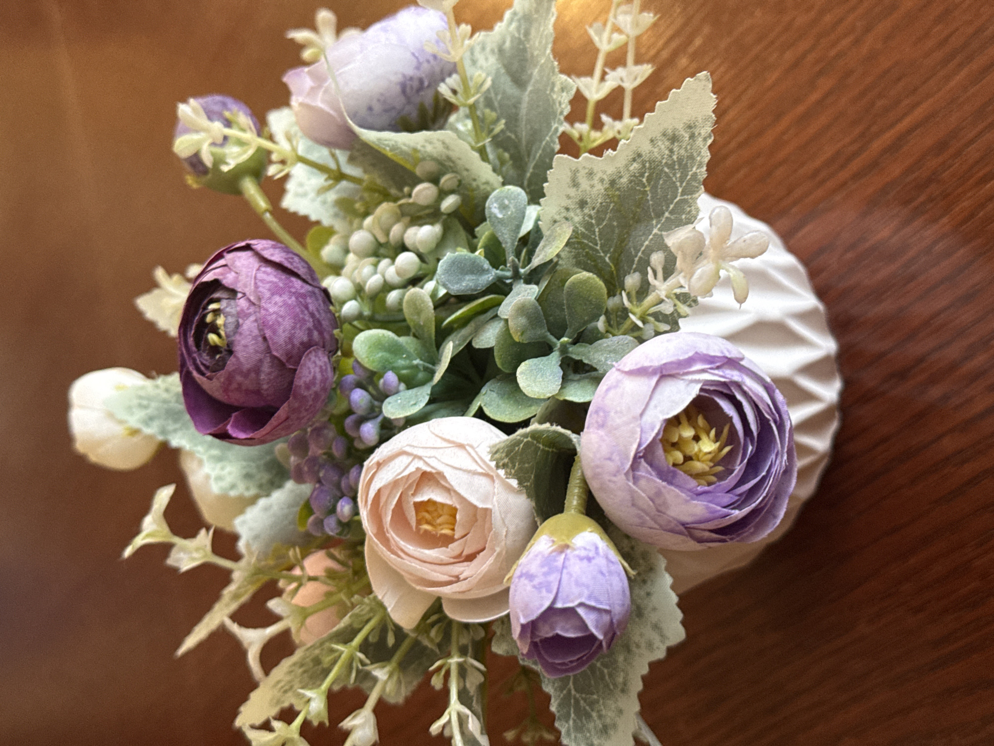 This is a close-up, overhead shot of a floral arrangement in a white, textured vase. The arrangement features several artificial flowers and greenery, primarily in shades of purple, pink, and white, with hints of light green and dusty grey foliage.
Prominent flowers include a deep purple peony-like bloom on the left, a soft pink peony-like bloom in the center, and a lighter purple flower to the right. There are also smaller buds and clusters of what appear to be small white berries or flowers interspersed throughout. The foliage is a mix of soft, dusty green leaves and some more delicate, sprig-like greenery with tiny white flower accents.
The vase itself is white with a diamond-like textured pattern. It is positioned on a dark brown, wooden surface, likely a table. The lighting suggests a warm, indoor environment, with a soft glow illuminating the top right portion of the image, hinting at a light source from that direction. There is no visible text in the image. The scene is static, with no activity taking place beyond the arrangement of the flowers. The overall impression is one of gentle beauty and crafted decoration.