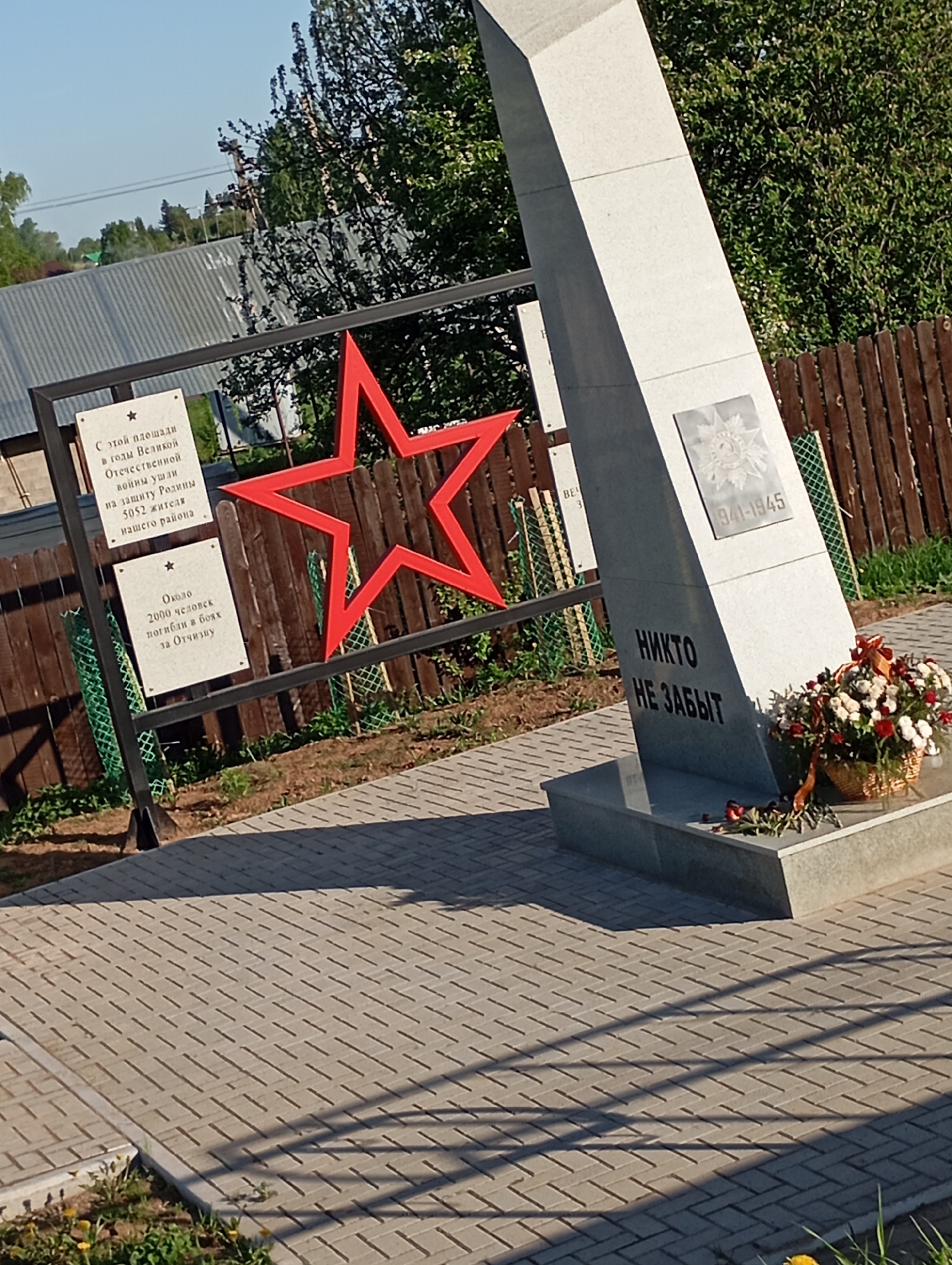 The image shows a memorial dedicated to those who fought in the Great Patriotic War (World War II). The memorial features a gray stone obelisk with the inscription "НИКТО НЕ ЗАБЫТ" ("No one is forgotten") in Cyrillic letters. Above the inscription is a plaque with a Soviet star and the dates "1941-1945". A basket of flowers sits at the base of the obelisk.
Next to the obelisk is a black metal frame with a large red star. Two plaques are attached to the frame. The top plaque reads: "С этой площади в годы Великой Отечественной войны ушли на защиту Родины 5052 жителя нашего района" (From this square, in the years of the Great Patriotic War, 5052 residents of our region went to defend the Motherland). The bottom plaque reads: "Около 2000 человек погибли в боях за Отчизну" (About 2000 people died in battles for the Fatherland).
The memorial is located in an outdoor area paved with rectangular gray pavers. A wooden fence is visible in the background, along with trees and a building. The memorial is bathed in sunlight. The location is in a city called Unknown, Unknown.