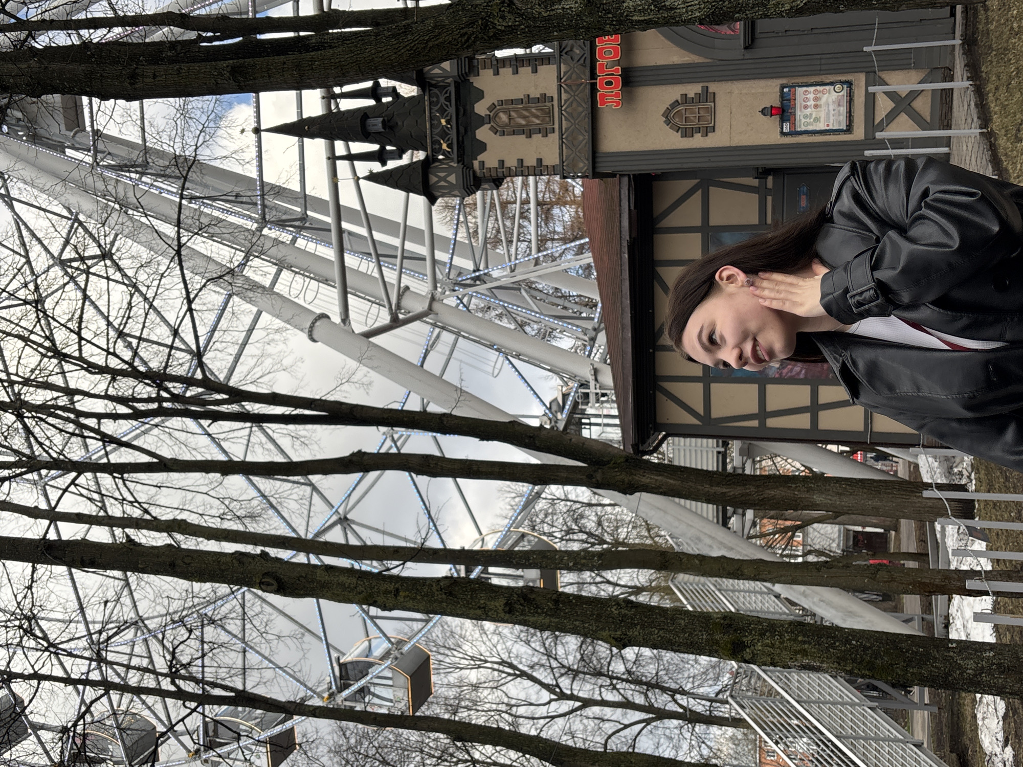 The image depicts a woman in a black leather jacket standing in front of a Ferris wheel, which is partially obscured by bare trees. Behind the woman and to the right is a building with intricate architectural details, including a tower-like structure and signage in Russian. The Ferris wheel is white with enclosed cabins. The setting appears to be outdoors in a park-like area. Based on the signage in Russian, it is likely this image was taken in the city of Perm, Russia.