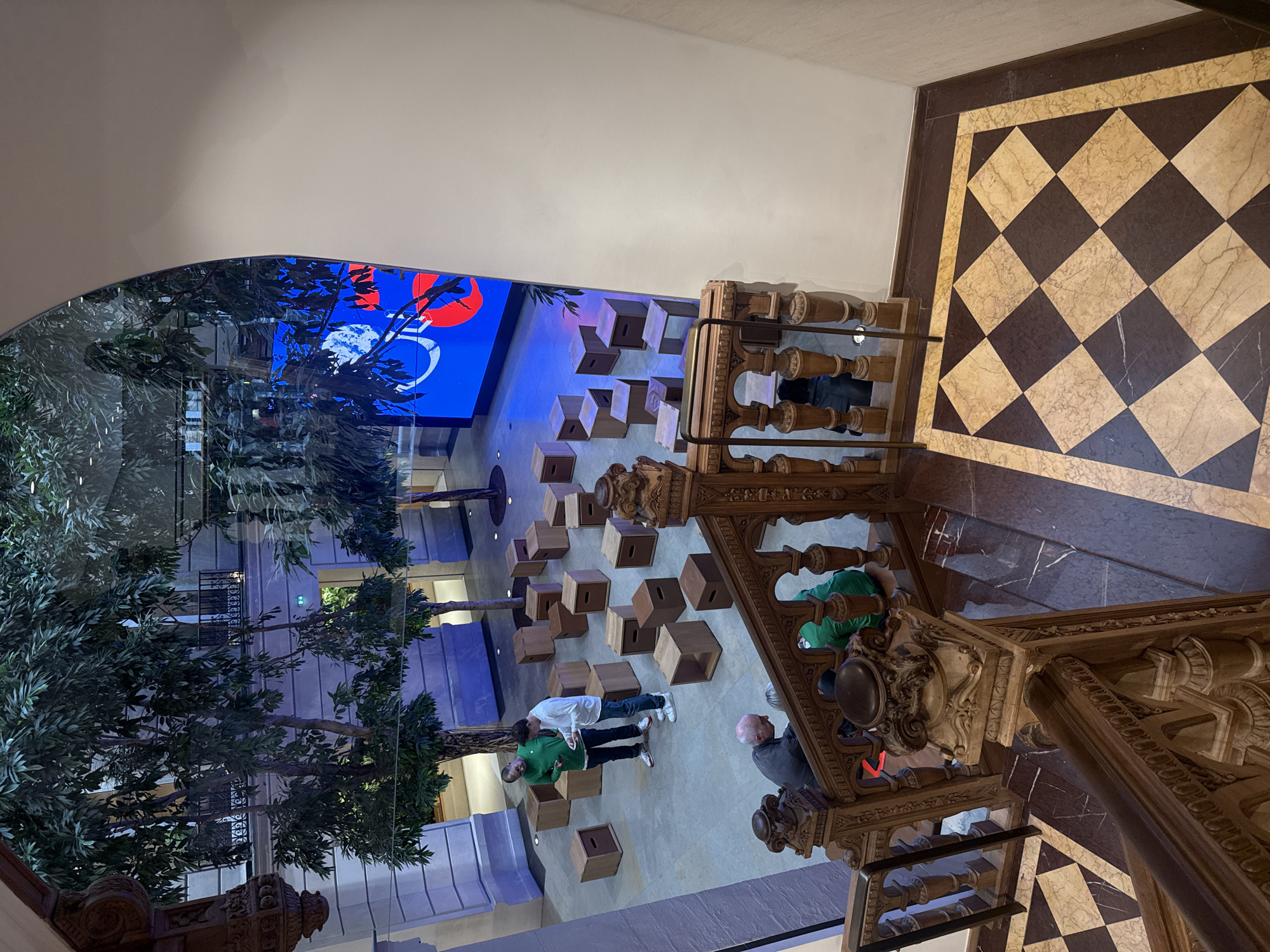 The photograph depicts an interior view of a building in Paris, France, seen from an elevated perspective, possibly from a staircase. The location appears to be a public space or atrium.
In the foreground, ornate wooden banisters and a staircase are visible, with a decorative, baroque style. The floor is a checkerboard pattern of dark and light squares.
Below, in the main atrium area, several wooden boxes are scattered on the floor, arranged somewhat irregularly. Two men are standing among the boxes, one wearing a green shirt and the other a white shirt. An older gentleman stands close to the staircase banister, closer to the camera.
In the background, there's a large blue screen with a red and white design. Trees are incorporated into the indoor space, adding a natural element to the architectural setting. Balconies are seen on the surrounding levels.