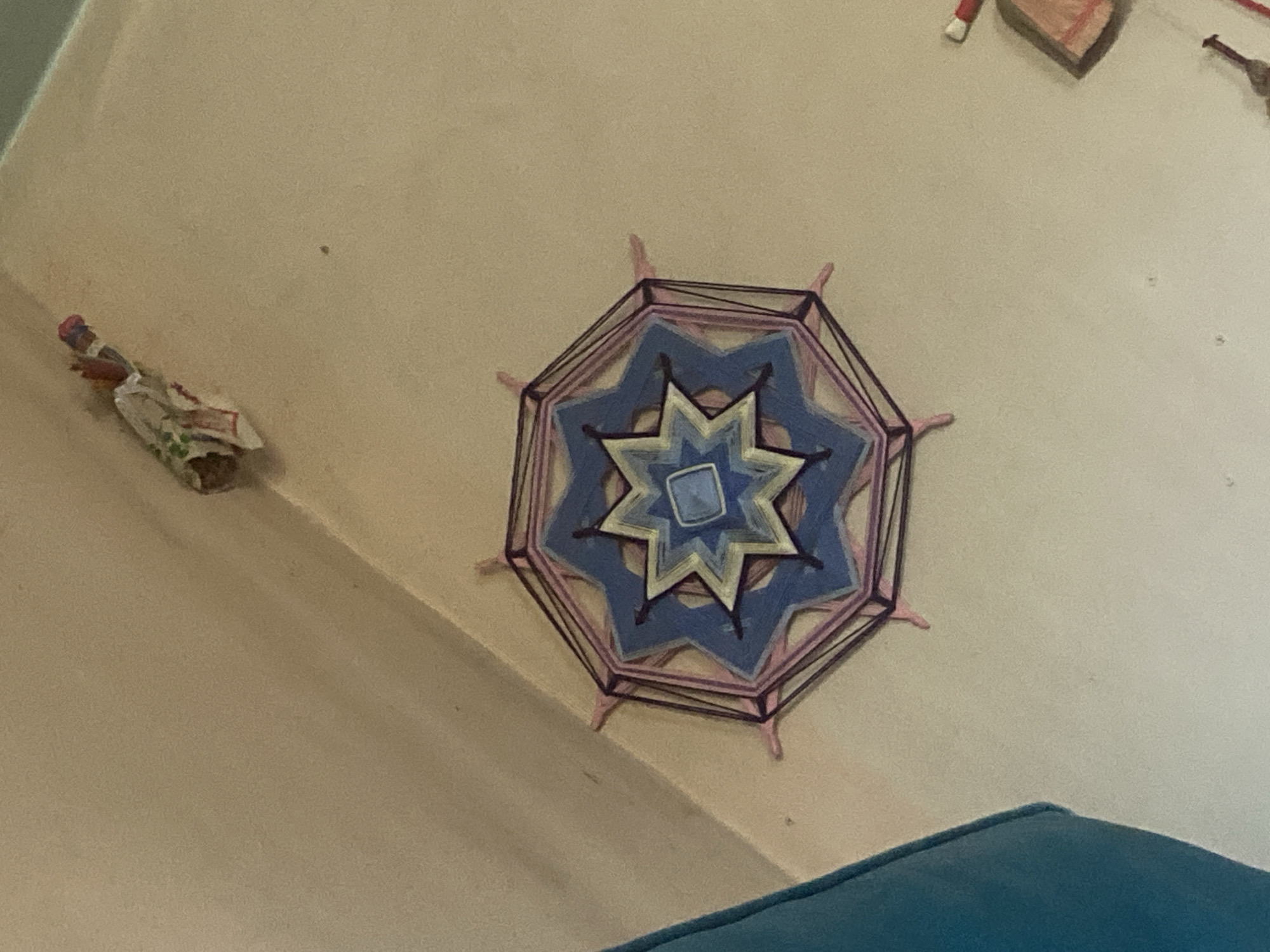 An octagonal woven mandala with intricate geometric patterns in shades of blue, white, and purple is displayed on a light beige wall. The mandala has eight pointed protrusions extending outwards, decorated with pink yarn. To the left of the mandala, a small, decorative figure resembling a doll or effigy, wrapped in white and red fabric, is positioned on the wall. In the upper right corner, a sliver of a wooden object, possibly part of a larger decorative piece, is visible. The wall appears to be a muted, neutral tone, possibly cream or light beige. The foreground shows a portion of a teal-colored surface, likely a piece of furniture, like a couch or chair. There is no visible indication of the location being in Черновая, Russia; the description is based solely on the visual elements present in the image.