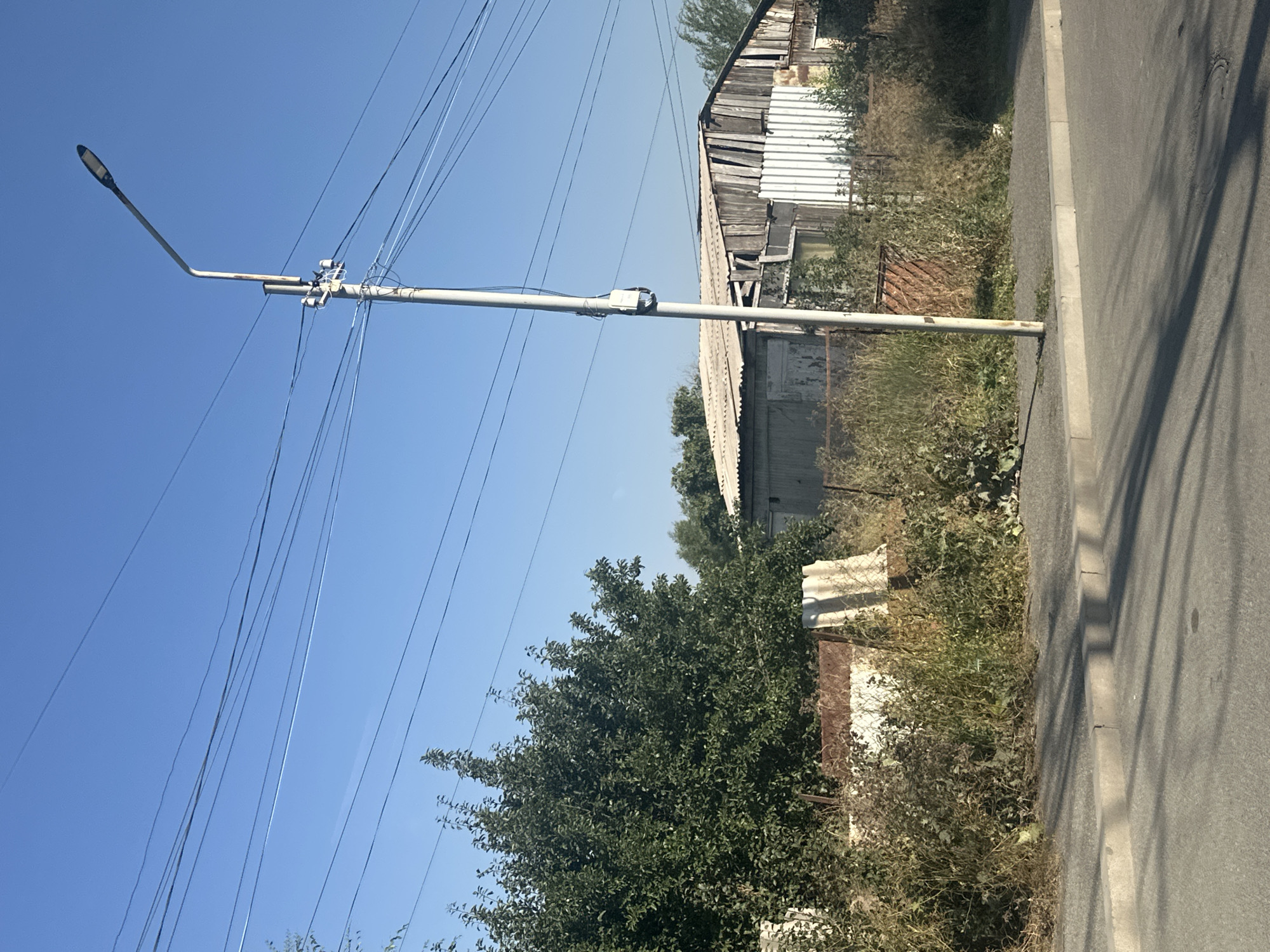 The image depicts a street scene in Gyumri, Armenia, under a clear blue sky. A tall, weathered streetlamp stands prominently on the right side of the frame, its arm extending to the upper right, casting a shadow on the asphalt road in the foreground. Multiple overhead wires crisscross the sky above the lamp and continue across the frame.
To the left of the streetlamp, a dense, green tree is visible, its leaves suggesting a summer or late spring season. Behind the tree and the streetlamp, a corrugated metal structure, possibly a shed or garage, is partially obscured by overgrown vegetation and rusted metal fencing. The building's roof appears to be made of the same corrugated material, some sections of which are damaged or peeling. Another, smaller structure with a light-colored corrugated roof is visible to the left of the main building.
The foreground consists of a paved road with a concrete curb and sidewalk. Shadows from the streetlamp and unseen objects are cast on the road surface. The overall impression is one of a slightly neglected, but functional, urban or suburban area. There are no people or animals visible in the image.