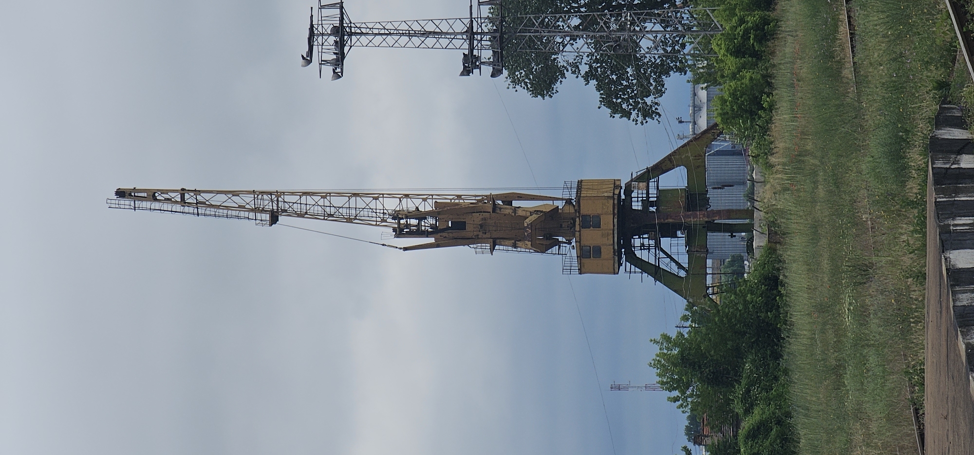 The image depicts an industrial scene in Svishtov, Bulgaria, featuring a large, weathered yellow gantry crane prominently in the foreground. The crane's metal structure is complex, with a tall boom extending upwards and a wider base supported by triangular legs. The main body of the crane has a box-like structure with windows. To the right of the crane, another metallic structure with what appear to be lights or speakers is visible, partially obscured by trees. The surrounding environment is overgrown with tall green grass and various bushes and trees. In the background, industrial-looking buildings or silos can be seen, along with what appears to be a radio tower in the distance. The sky is overcast, contributing to a somber atmosphere. The scene suggests an abandoned or disused industrial area, likely part of the port or a similar facility in Svishtov. There are no people or animals visible in the image.