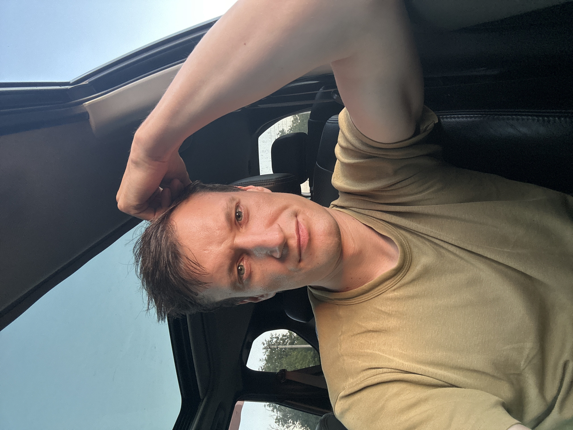 A man sits in the driver's seat of a car, his right arm raised and resting on his head. He is looking directly at the camera, with a neutral expression. The man is wearing a light brown t-shirt. The car's sunroof is open, revealing a clear blue sky. The background shows trees and foliage visible through the car's windows, suggesting an outdoor setting. This image does not provide enough information to determine if it was taken in the city of Makiv, Ukraine.