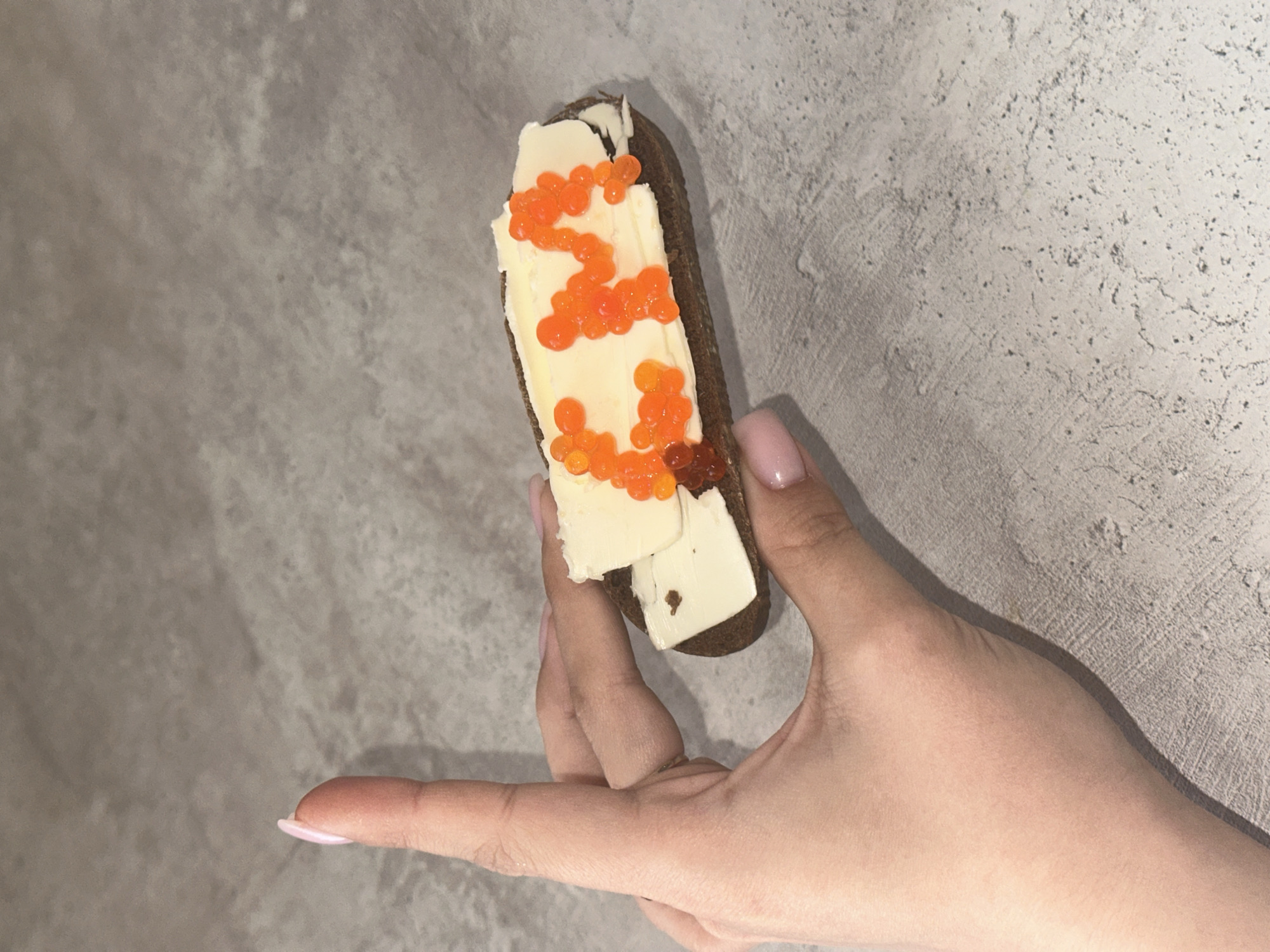 A hand holds a piece of dark bread topped with butter and orange caviar against a gray textured background. The person has manicured nails with pink polish. The location is not discernible from the image.