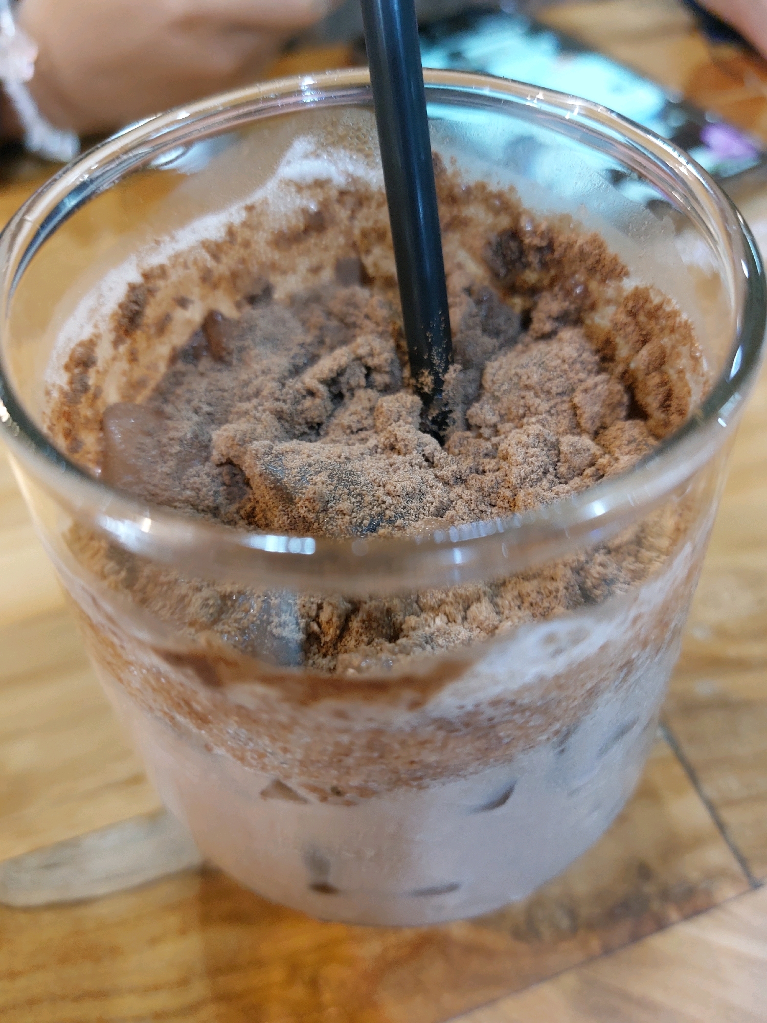 A close-up shot shows a glass of iced coffee with a black straw. The drink is topped with a generous layer of chocolate powder. The glass is sweating, indicating it is cold, and is sitting on a wooden table. In the blurred background, a person's arm and hand are visible, along with some other out-of-focus objects. This scene could be found in various cafes or restaurants in Tarakan, Indonesia.