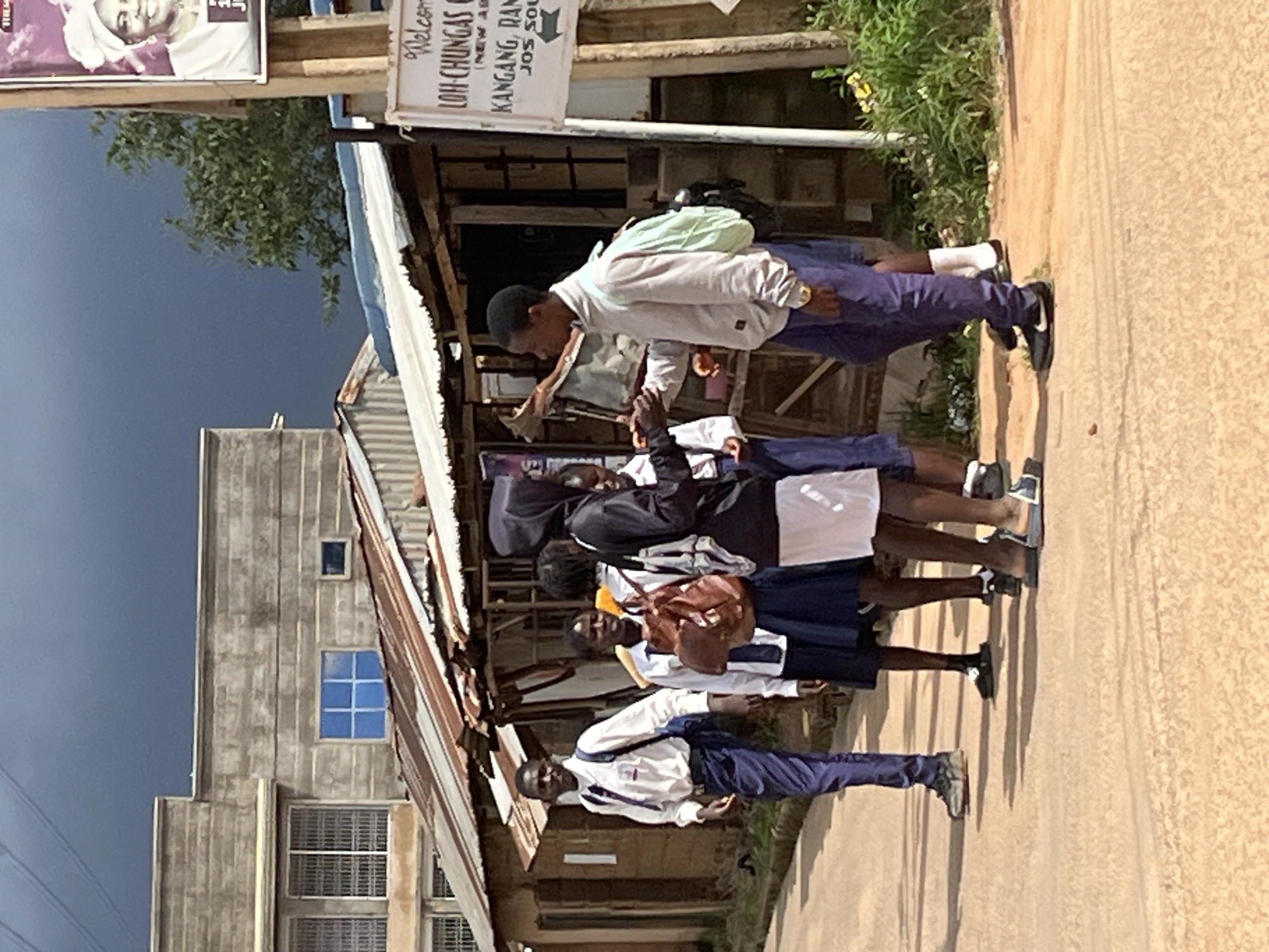 A group of five young people, likely students, are standing in a line on a dirt road in what appears to be a rural or semi-urban area. They are wearing school uniforms, consisting of white shirts and blue trousers or skirts. One of the individuals in the front is wearing a white shirt and blue trousers, and has a black backpack on their shoulders. The person behind them is wearing a white shirt and dark skirt, with a brown backpack. The other students also appear to be in uniform.
The scene suggests a normal day, possibly before or after school. The lighting indicates it is daytime, with shadows cast on the road, suggesting it could be mid-morning or afternoon. The weather seems clear, with a bright sky visible in the background, though there is a hint of darker clouds towards the left side of the image.
Behind the group, there is a building with a corrugated iron roof and some wooden structures, suggesting a modest commercial area or possibly a school or community center. A sign with text is visible on a wooden post. The text on the sign is in English and appears to be directional, with "Welcome" at the top, followed by place names like "LOH-CHUNGAS" and "KANGANG, RANT," and "JOS SOUTH." The building to the left is a more substantial structure with windows, some of which are barred, and appears to be made of concrete.
The overall impression is one of everyday life in a Nigerian community. The individuals appear neutral in expression, focused on whatever is happening off-camera or on their way somewhere.