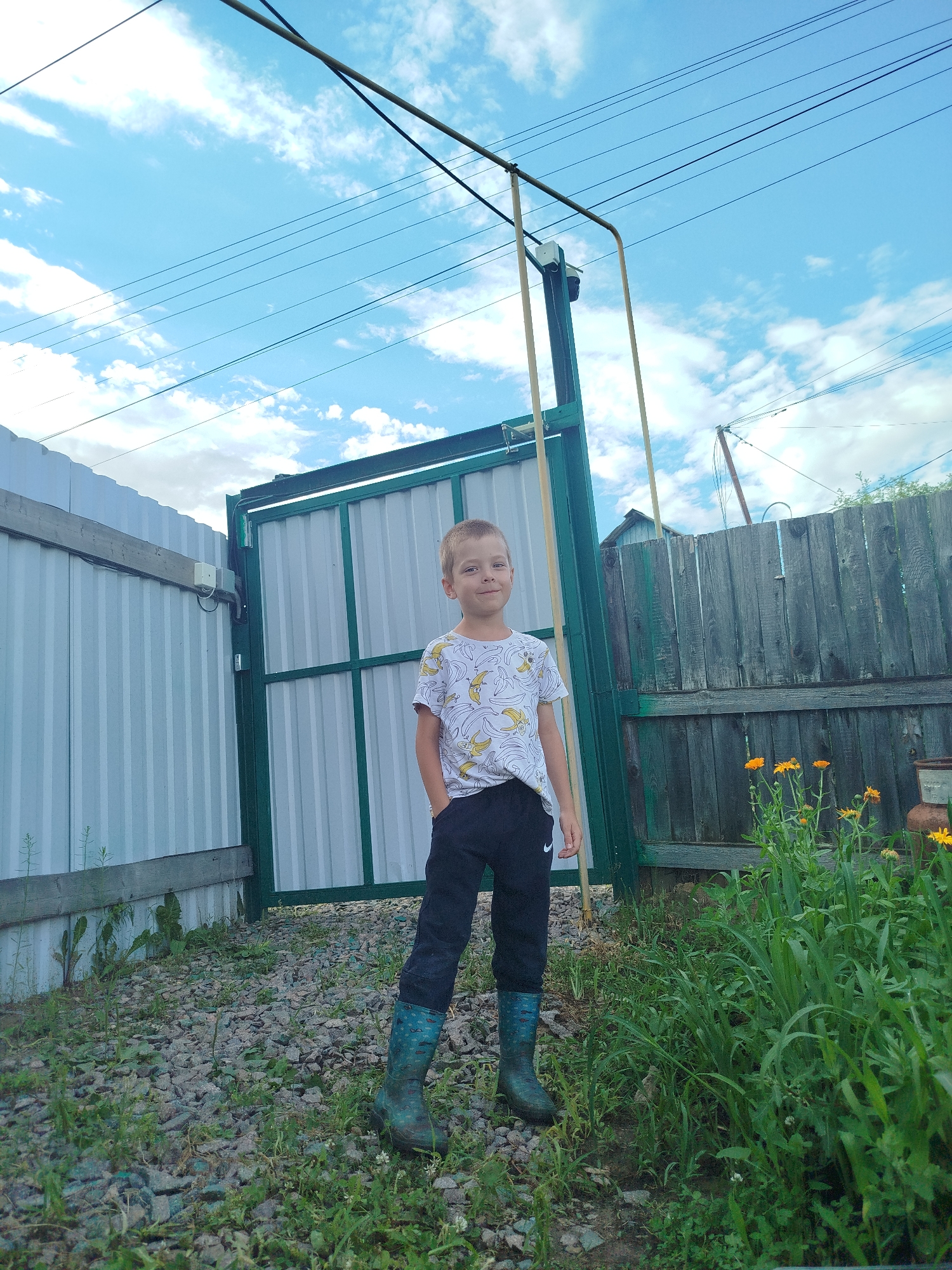 A young boy stands in the foreground of an outdoor scene, facing the camera with his left hand in his pocket. He is wearing a white t-shirt with a banana print, black pants, and blue patterned rubber boots. Behind him is a green metal gate, flanked by a white corrugated metal fence on the left and a weathered wooden fence on the right. The ground is covered in gravel and some sparse grass. In the background, there are power lines against a blue sky with scattered clouds. Orange flowers are visible near the wooden fence on the right. The setting appears to be a backyard or yard area.