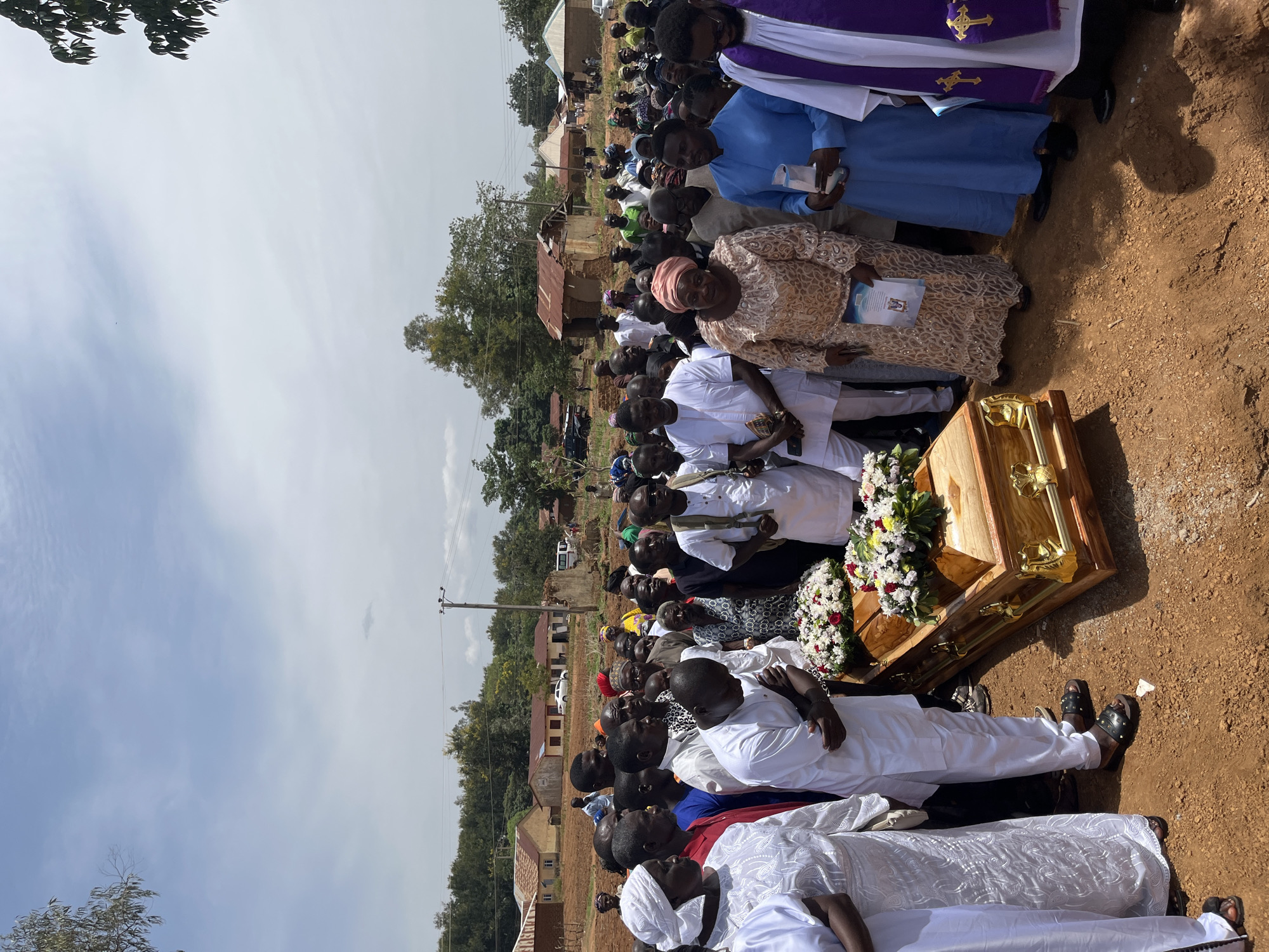 A large group of people is gathered outdoors, likely in Dogon Dinia, Nigeria, near a wood coffin decorated with flowers. Many individuals are dressed in white, while others wear a variety of colors and patterns. A priest stands near the coffin wearing a purple and white robe with a cross symbol. The environment includes dirt ground, trees, houses, and power lines, suggesting a rural setting.