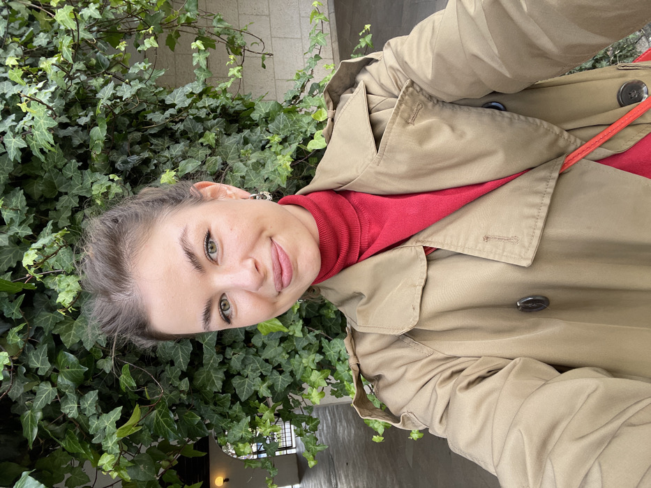A female individual is centered in a selfie-style, eye-level photograph, located within the city of Budva, Montenegro. She possesses light skin, green eyes, and brown hair secured in an updo. Her attire consists of a red turtleneck sweater layered under a light tan double-breasted trench coat, with a partial view of a red strap visible across her lower chest. Small, silver-toned hoop earrings are visible in both ears. She maintains direct eye contact with the camera and displays a subtle, closed-lip smile. The background immediately behind her and to her right is dominated by a wall densely covered with vibrant green ivy. To her left, an arched, off-white architectural structure, consistent with a covered walkway or arcade, recedes into the background, featuring an illuminated light fixture. Beyond this structure, a metal barrier and distant green foliage are present.