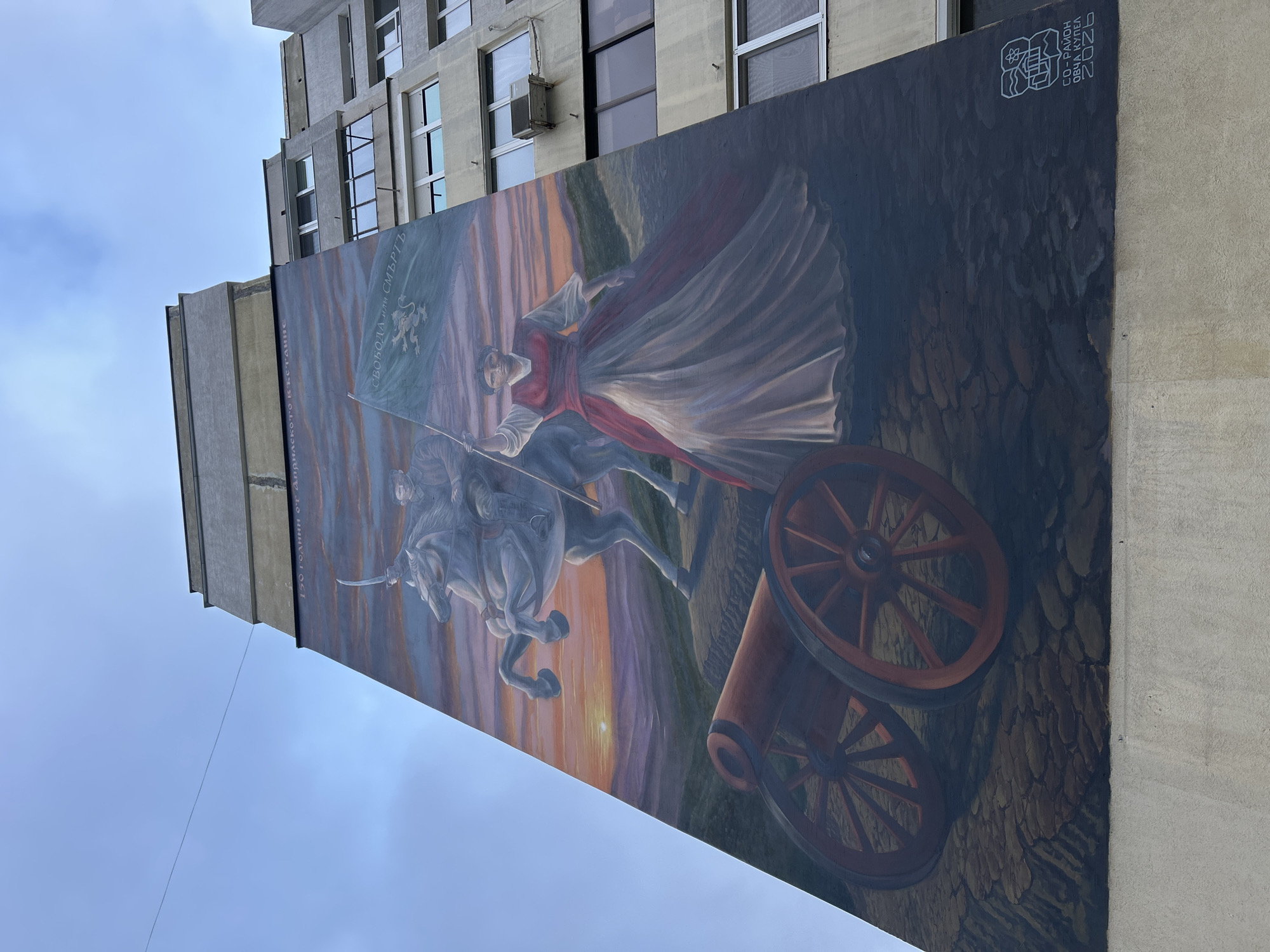 A low-angle shot depicts a large mural painted on the side of a concrete building. The mural features an image of a person riding a horse while holding a flag. Another person walks alongside the horse, and a cannon is visible. A cityscape is painted in the background of the mural. At the top-right of the mural, a white logo with text is visible. Above the mural is a concrete building with several windows. A blue sky is visible in the background. Location is Unknown, Unknown.