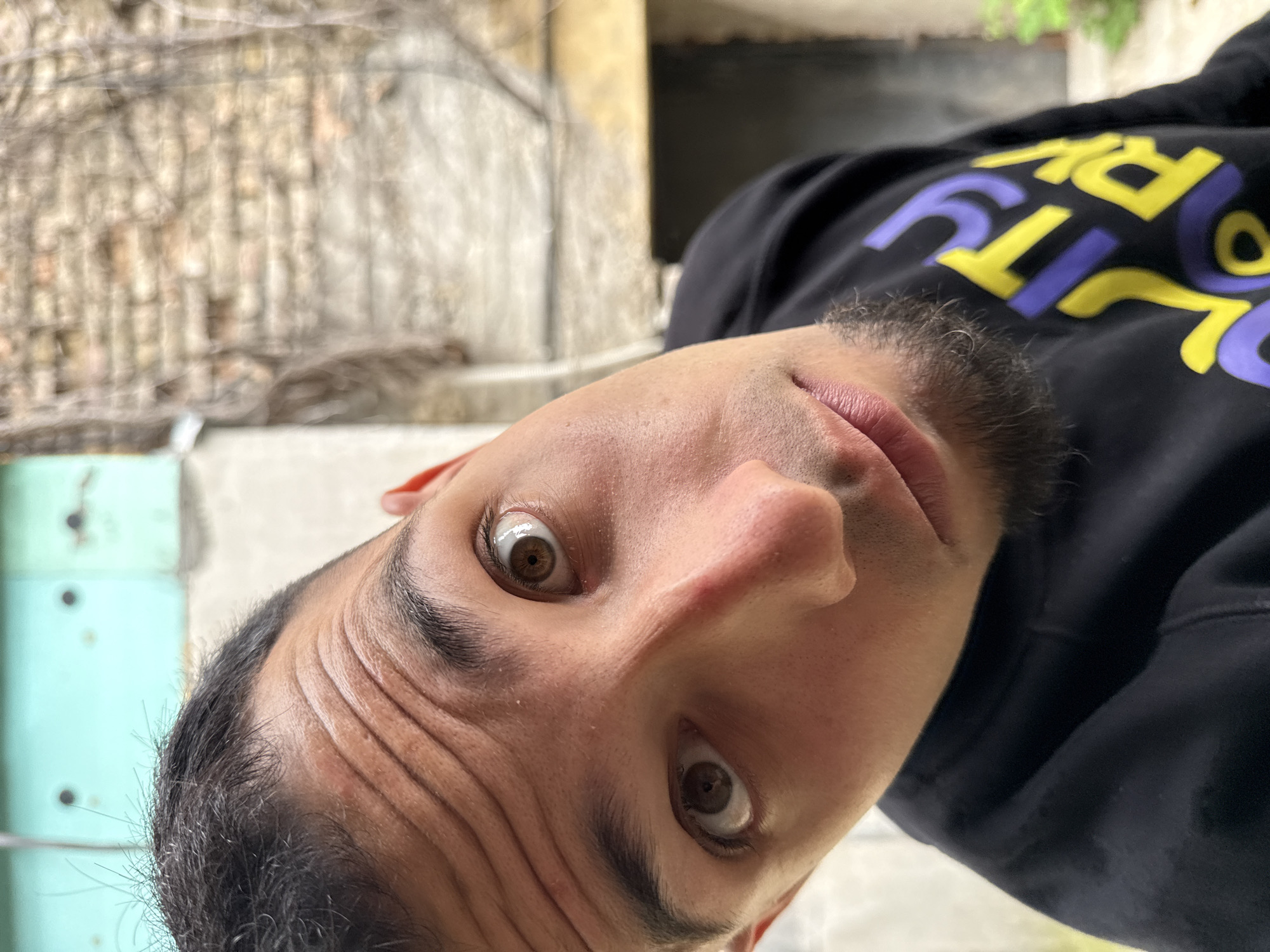 A close-up shot of a man's face is presented at an unconventional angle, rotated counter-clockwise. The man gazes directly into the camera with wide eyes, showcasing brown irises. The face displays prominent forehead wrinkles and a neatly trimmed, dark beard. The man wears a black hoodie with a design featuring the word "DIRTY" in yellow and purple lettering. In the blurred background, hints of a building exterior are visible on the left, constructed from light-colored, textured stone. The right side of the background includes blurry green foliage. Based on contextual information, the location is the city of Shumen, Bulgaria.