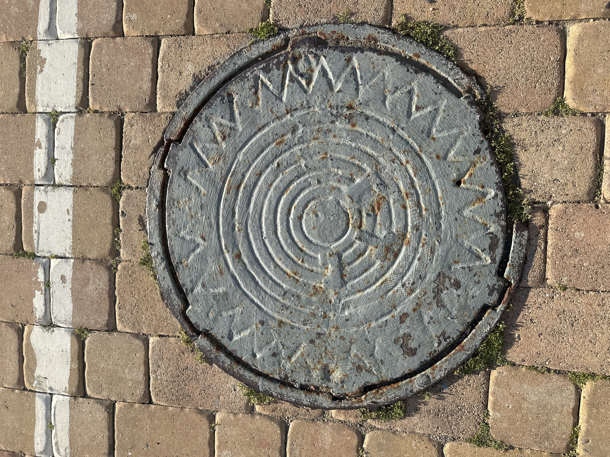 The image shows a circular, weathered metal manhole cover set into a brick sidewalk. The manhole cover has a concentric circular pattern and a zigzag design around its outer edge. Some patches of green moss and grass are growing around the edges of the manhole cover and in the gaps between the bricks. The sidewalk is composed of rectangular, reddish-brown bricks, with some sections of white paint markings visible at the top of the image. The lighting suggests it is daytime, casting shadows on the textured surfaces. There are no people or animals present in the scene. The overall impression is one of an urban streetscape, possibly in a public walkway or square.