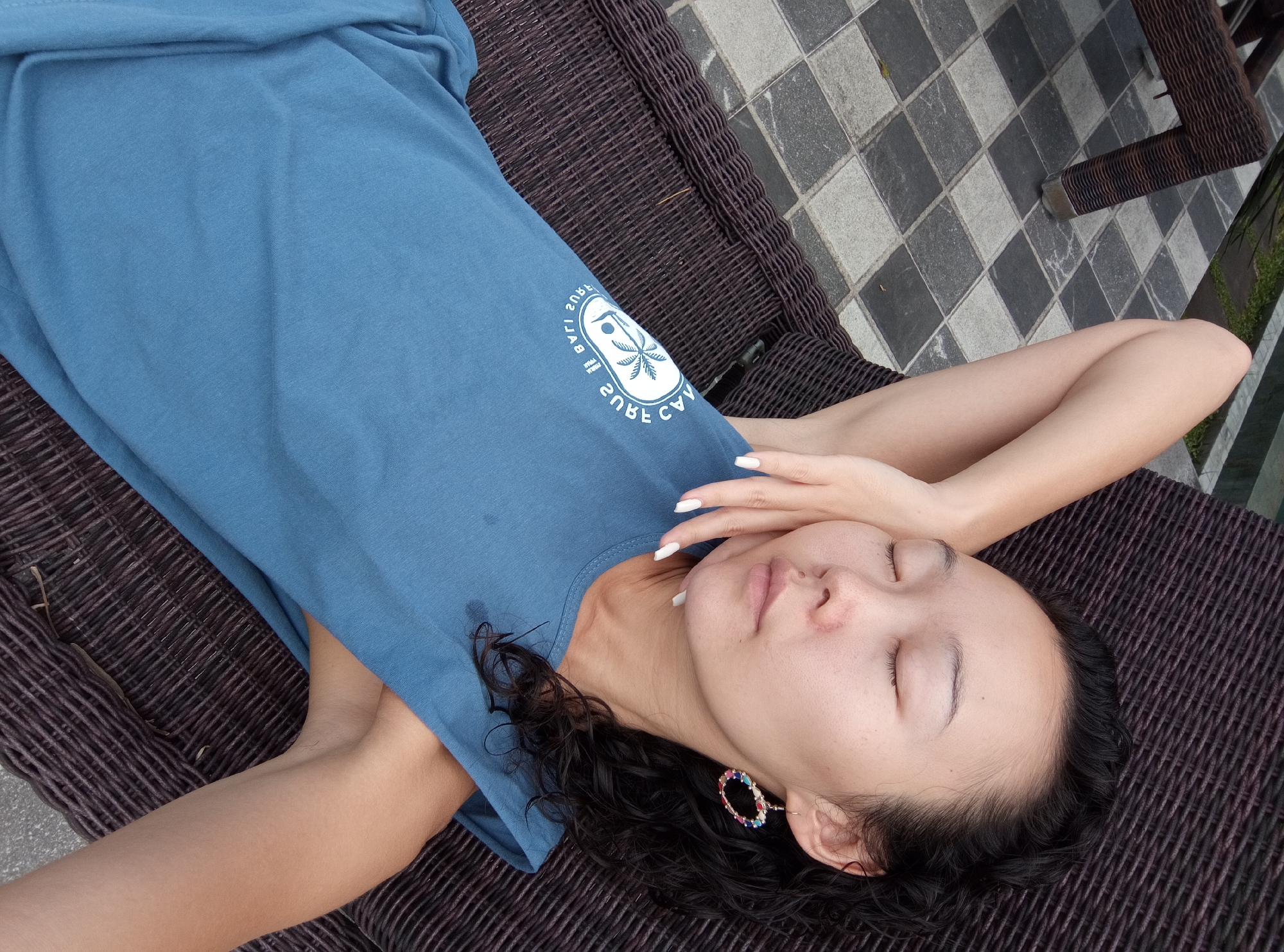 A young woman with dark hair and closed eyes is lying on a dark brown wicker lounge chair. She is wearing a blue tank top with a white circular graphic on the front that depicts a palm tree and the text "CAU 2 BALI SURF". Her right arm is bent and resting on her forehead, and her left arm is bent and resting on her chest. She is wearing a colorful hoop earring in her left ear and her fingernails are painted white. The lounge chair is situated on a tiled patio with a checkered pattern of gray and black squares. A portion of a dark brown wicker chair leg is visible in the upper left corner of the image. The environment appears to be outdoors, possibly near a pool or a garden, given the presence of greenery in the background and the tiled patio. The lighting suggests it is daytime.