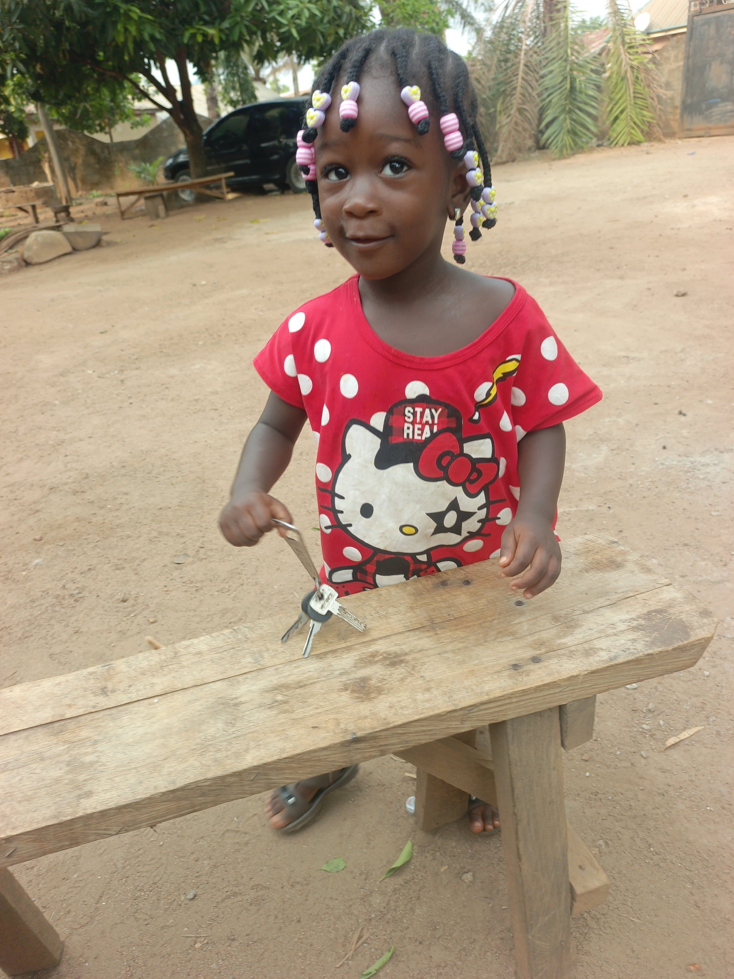 A young child stands behind a wooden bench on a sandy ground in Akwanga, Nigeria. The child, with dark skin and braided hair adorned with colorful beads, wears a red and white polka-dot shirt featuring a Hello Kitty graphic. The child's hands rest on the bench, holding a set of keys. The bench is a simple wooden structure with a plank seat and cross-braced legs. The ground is sandy and scattered with leaves. In the background, there is a car, trees, and other buildings. The sky is overcast.