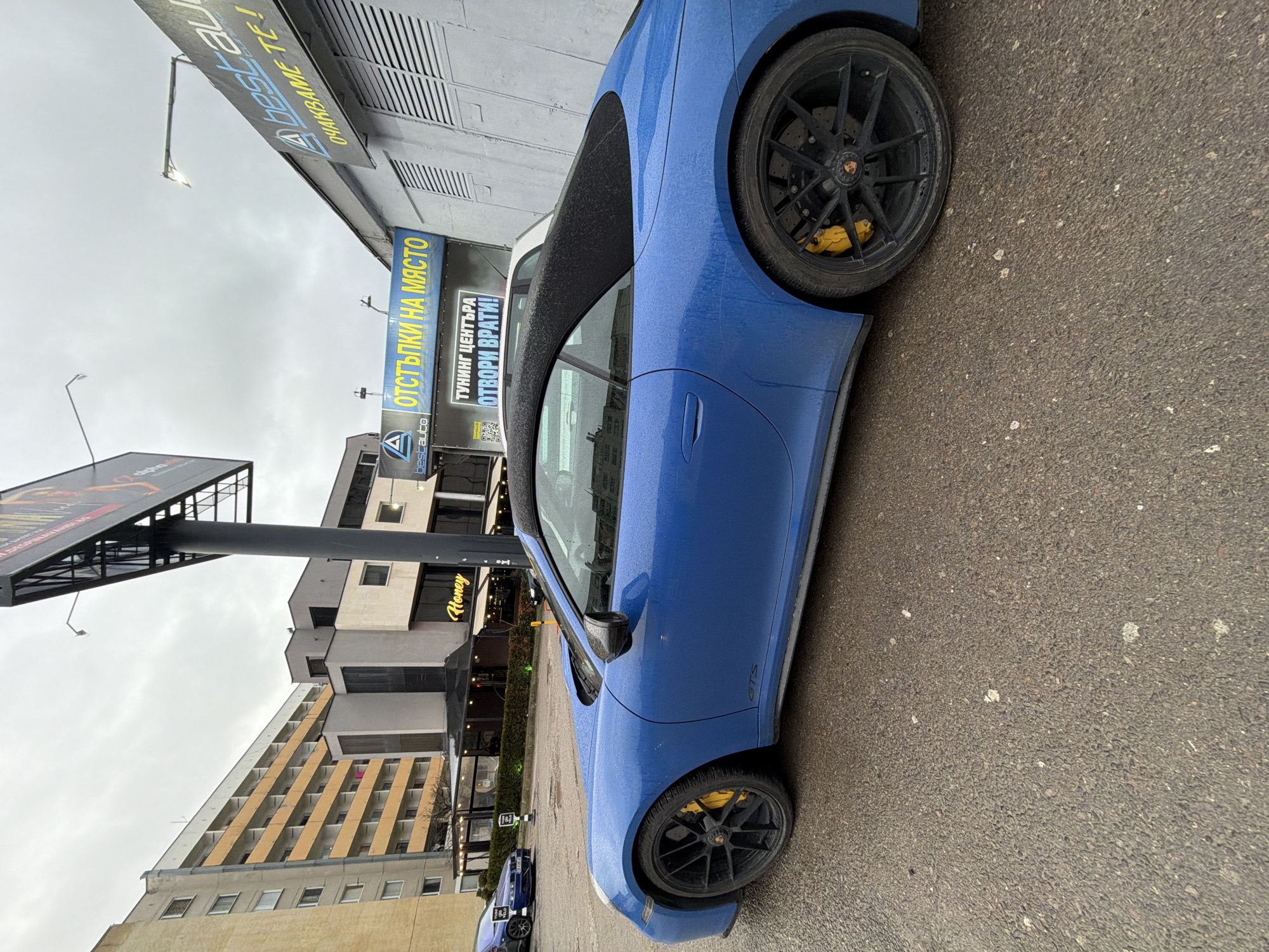 The image shows a blue Porsche 911 GTS parked on a street in Sofia, Bulgaria. The car is positioned on the right side of the frame, with a partially visible black soft top and black wheels with yellow brake calipers. A "GTS" badge is visible on the car's side. The background features several buildings, including a tall residential building with beige balconies and a building with a sign that reads "Honey." A large advertisement board is also present. There are commercial signs in Bulgarian on the buildings that read "ОТСТЪПКИ НА МЯСТО", "ТУНИНГ ЦЕНТЪРА ОТВОРИ ВРАТИ!", and "ОЧАКВАМЕ ТЕ!".