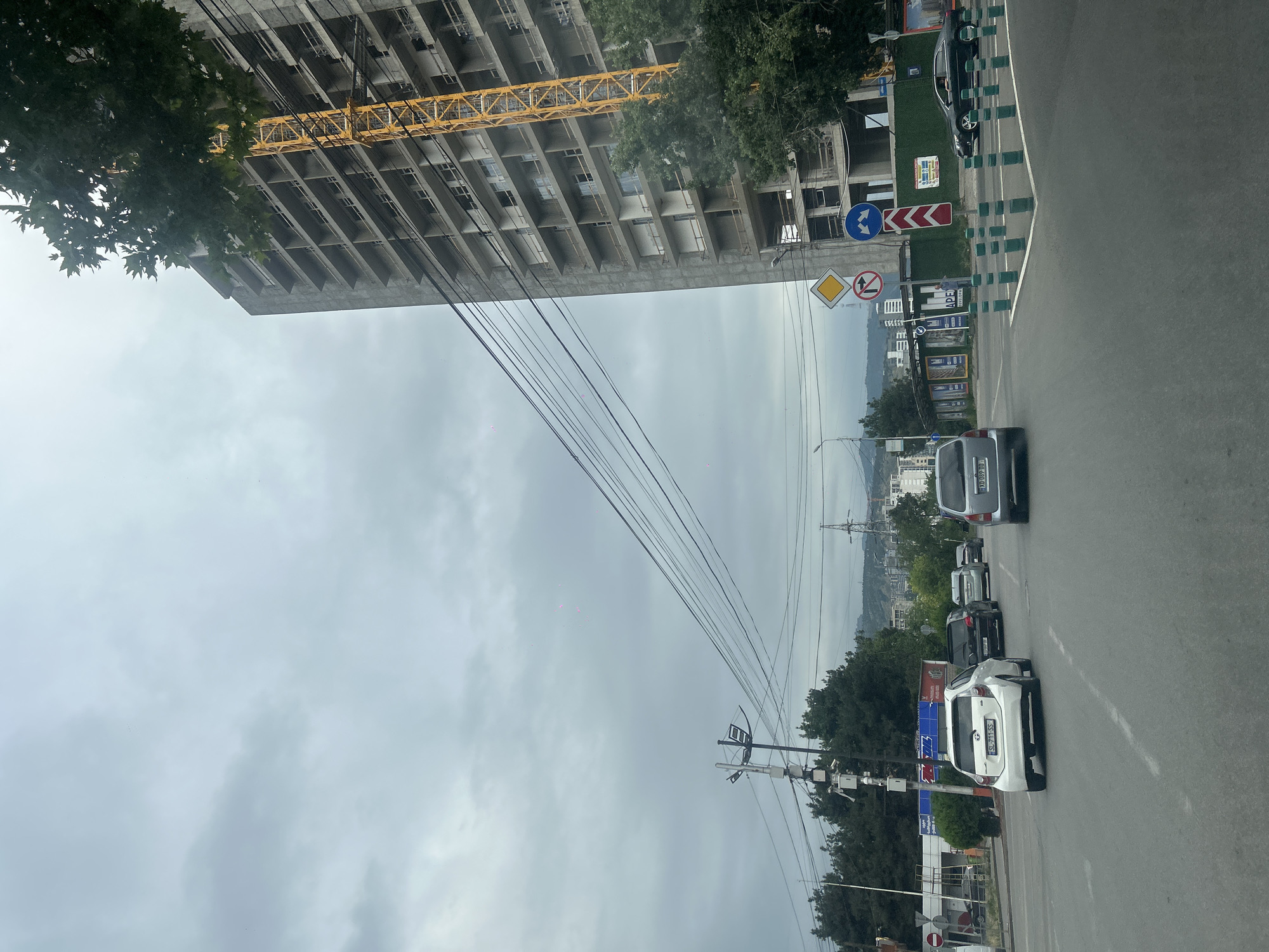 The image depicts a cloudy day scene on a street in Tbilisi, Georgia. A multi-story building under construction with a yellow crane is visible on the right side of the frame. Several cars are driving on the road, including a white hatchback in the foreground, followed by a dark SUV and other vehicles. Traffic signs, including a diamond-shaped yield sign and a directional sign indicating a turn, are present at an intersection. Utility poles with numerous wires run across the sky. Trees line the side of the road and surrounding areas. In the background, other buildings and hills are visible.