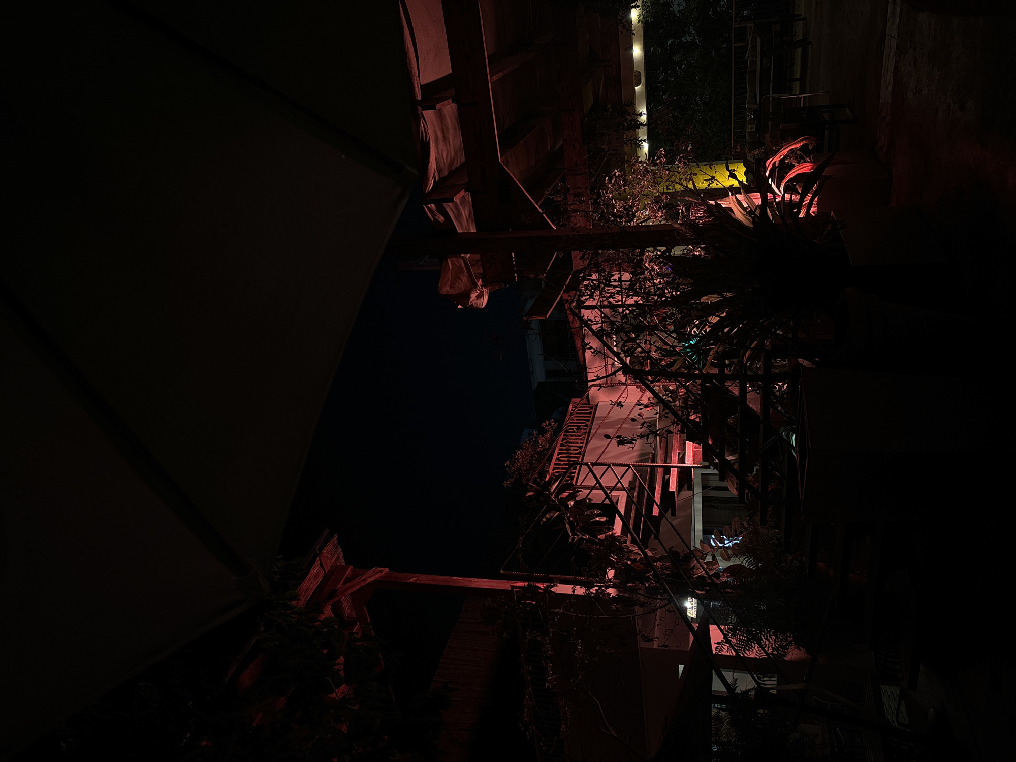 The image depicts an outdoor seating area at night, illuminated by red and yellow lights. A large, dark awning or roof covers a portion of the scene. In the background, a set of stairs is visible, lit by red lights, leading up to a building with a sign that reads "PIONERE RESORT". Greenery, including potted plants and vines, is scattered throughout the area, with some foliage also illuminated by the red lights. Tables and chairs are visible in the dimly lit background, suggesting a restaurant or bar. The overall ambiance is dark and moody due to the nighttime setting and the colored lighting.