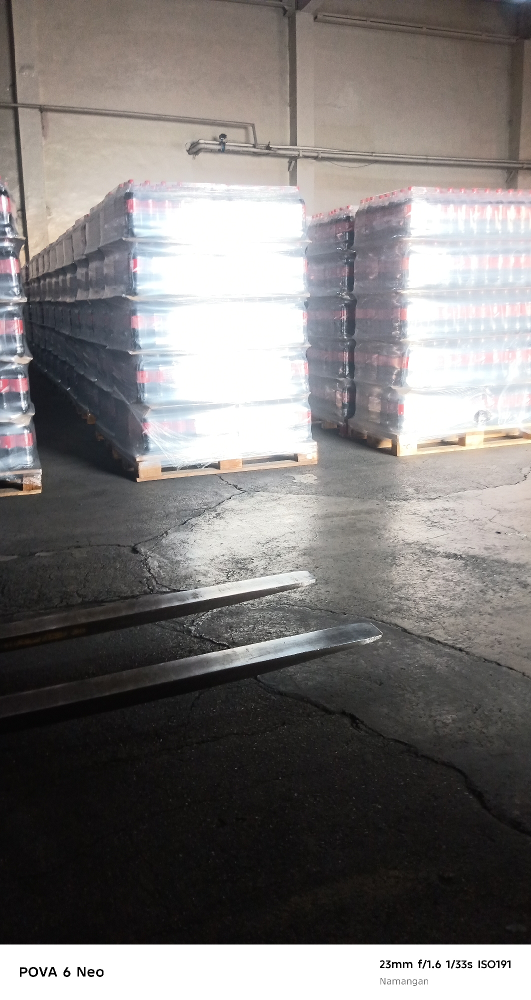 The image shows stacks of bottled drinks wrapped in plastic on wooden pallets in a warehouse. The floor is concrete with visible cracks. In the foreground, two metal fork truck tines are visible, partially submerged in shadow. The lighting suggests an indoor industrial environment. The location is identified as Namangan City, Uzbekistan.
