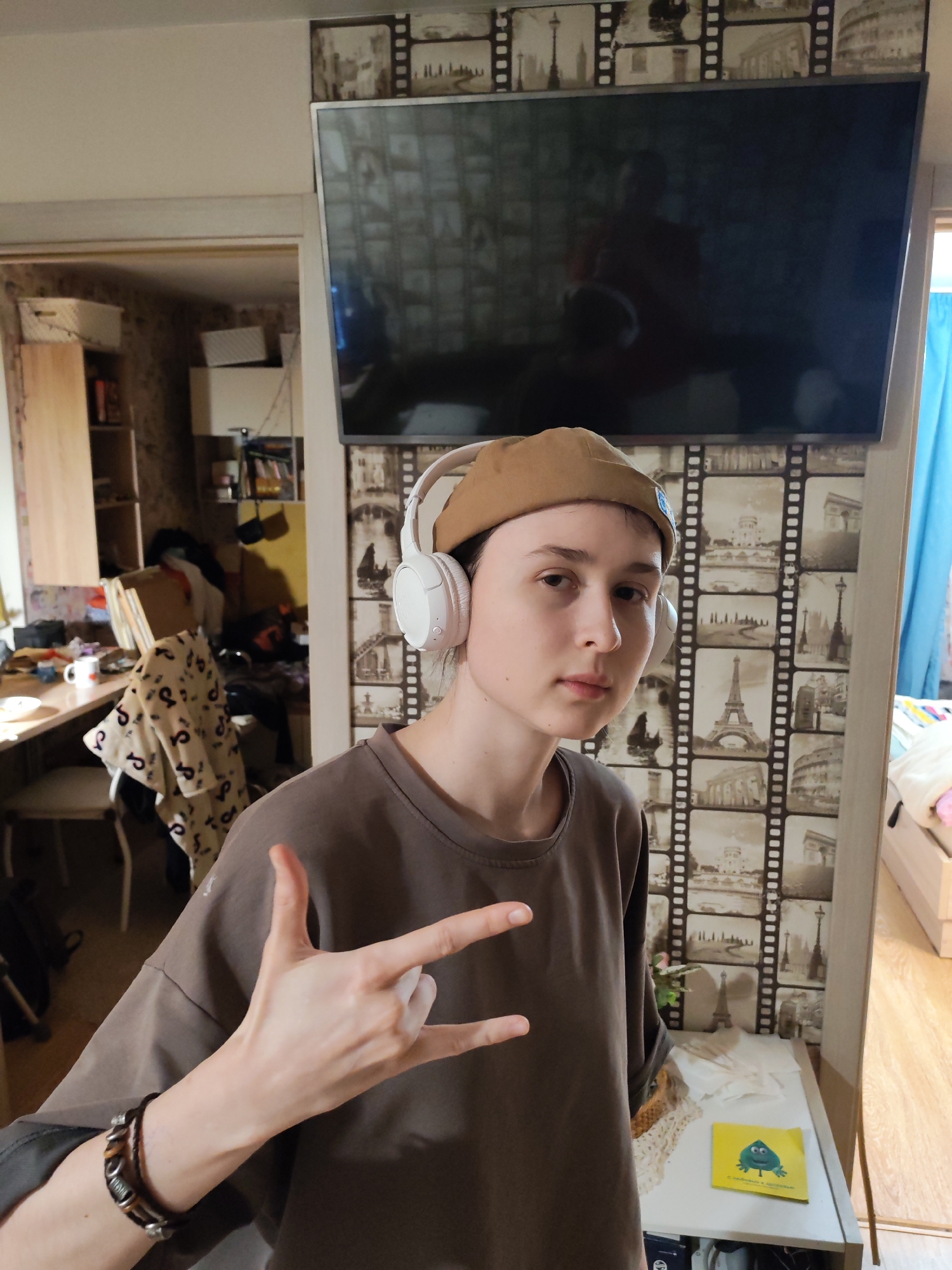 The scene depicts a young individual in an indoor residential setting in Chaikovsky, Russia. The individual, who appears to be an adolescent, stands centered in the frame, making a "rock on" hand gesture with their left hand, fingers extended. They wear a light brown flat cap or beret, white over-ear headphones, and a plain dark brown short-sleeved t-shirt. A multi-strand bracelet is visible on their left wrist.
The background wall behind the individual is covered with wallpaper featuring a repeating pattern of film strips containing sepia-toned images of various European landmarks, including the Eiffel Tower, the Colosseum, and the Arc de Triomphe. A large, dark-screened flat-screen television is mounted on this wall, reflecting an indistinct image of the room and potentially the photographer. Below the television, a white shelf holds a small, green artificial plant, several white folded papers, and a yellow booklet with a blue cartoon character on its cover. To the right of the television, a section of a light blue curtain is visible. The floor appears to be a light wood laminate.
To the left of the individual, an open doorway reveals a second room that appears somewhat cluttered. Visible items in this adjacent room include a wooden desk or table with a white mug, a chair draped with a patterned blanket, and shelving units containing books, boxes, and other miscellaneous items. The walls in this second room also feature wallpaper, distinct from the film strip pattern in the foreground.
