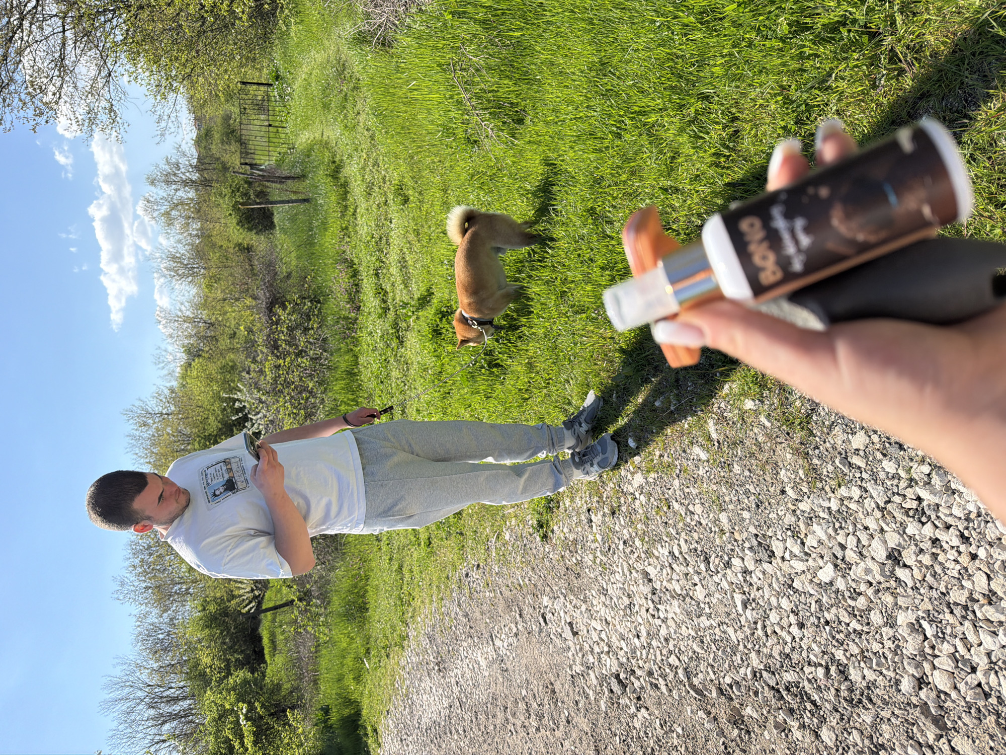The image depicts an outdoor scene in Priseltsi, Bulgaria. A man stands on a gravel path, looking at his phone. He wears a white t-shirt and grey sweatpants. To his right, a medium-sized, brown dog with a dark collar stands in a field of green grass. In the foreground, a hand holds a small spray bottle labeled "Bono." The background includes trees and a partially visible metal fence. The sky is partly cloudy.