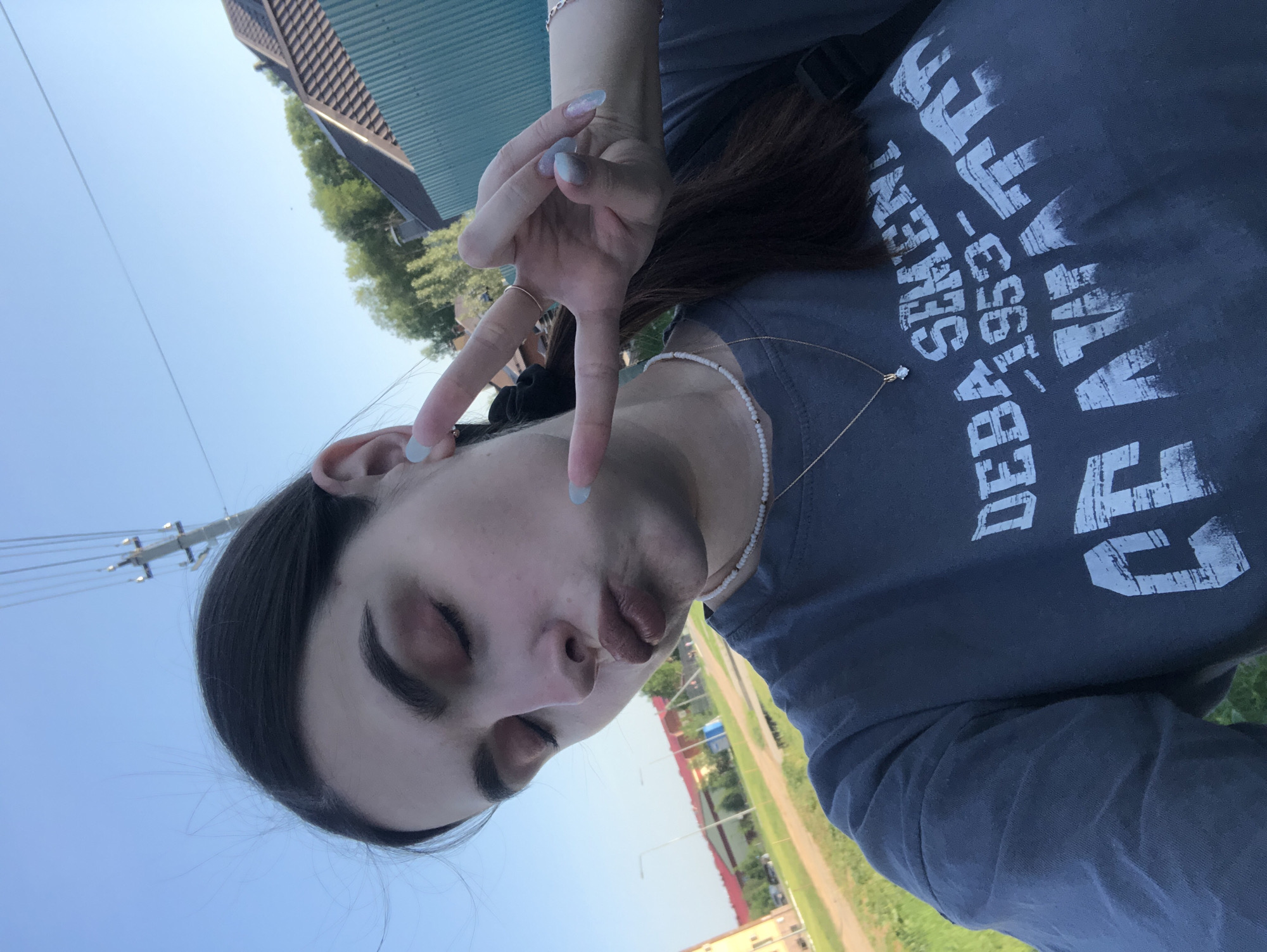 A young woman is facing the camera, tilted to the side. She is making a peace sign with her right hand. She wears a grey shirt with a faded graphic print. The background includes a street with buildings, trees, and a clear blue sky. It is possibly a street scene in Noviy, Russia.