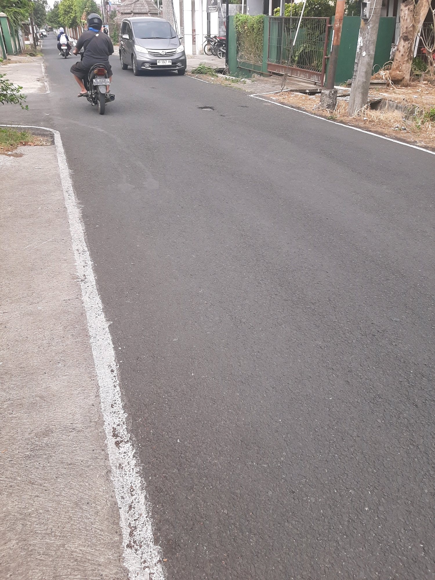 A street scene in Sumampir, Indonesia, shows a paved road with a concrete sidewalk on the left. Several motorcycles are visible, with one prominent motorcycle carrying a rider wearing a dark shirt, black pants, and a helmet, moving away from the camera. Another motorcycle is parked in the background on the right. A silver car with license plate "AD 1347 PB" is parked on the road. The environment includes houses with green fences and trees. The road surface is asphalt with a white line marking the edge of the sidewalk.