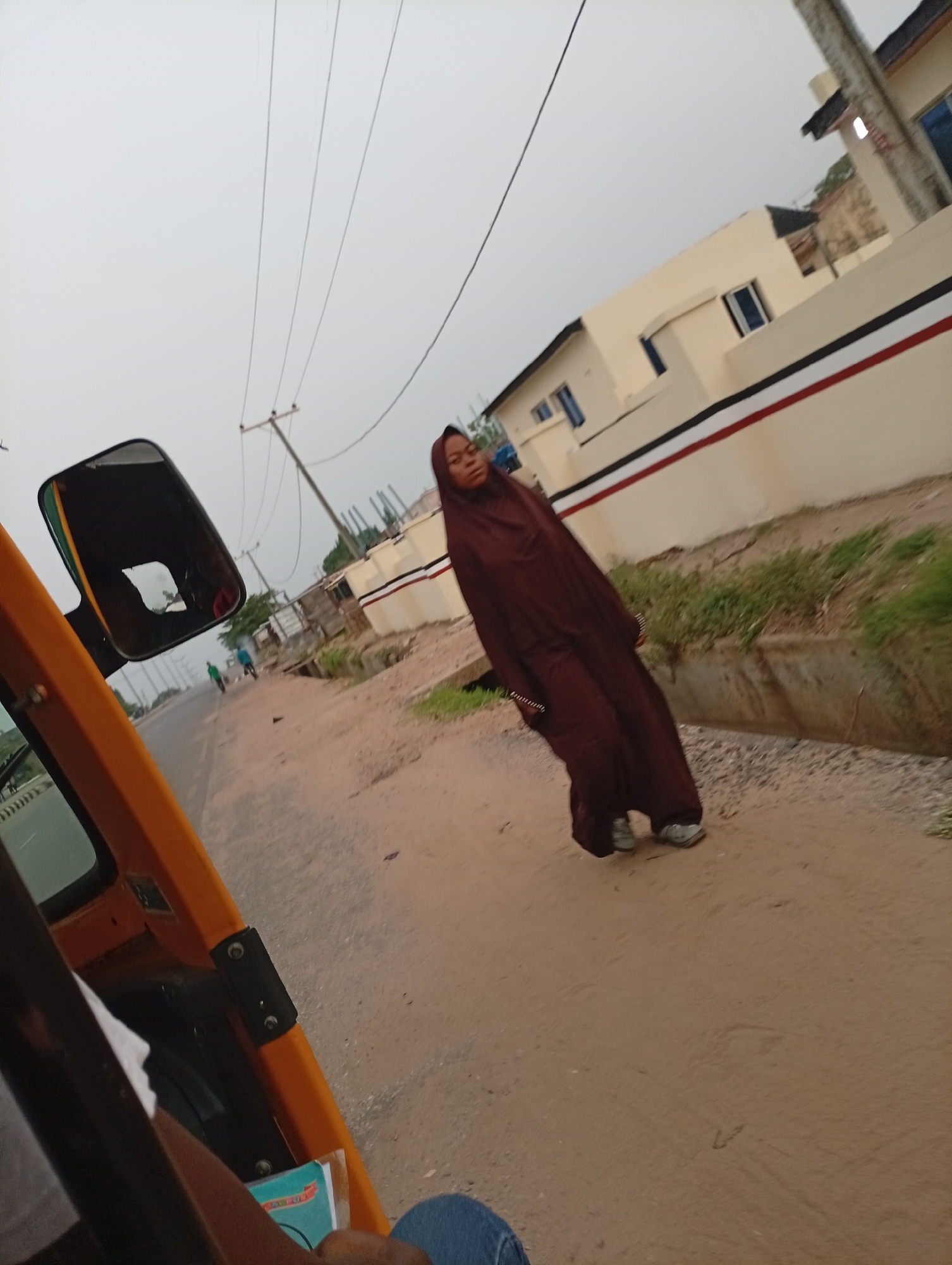 A woman wearing a dark brown abaya walks alongside a road in Adankolo, Nigeria. The road appears unpaved. A ditch runs along the side of the road. Behind the woman is a white building with a red and black stripe painted on its wall. Power lines extend into the sky. The perspective is from inside a yellow vehicle, likely a tricycle or taxi, with part of the dashboard and the driver's arm visible. Other pedestrians are seen in the distance. The sky is overcast.