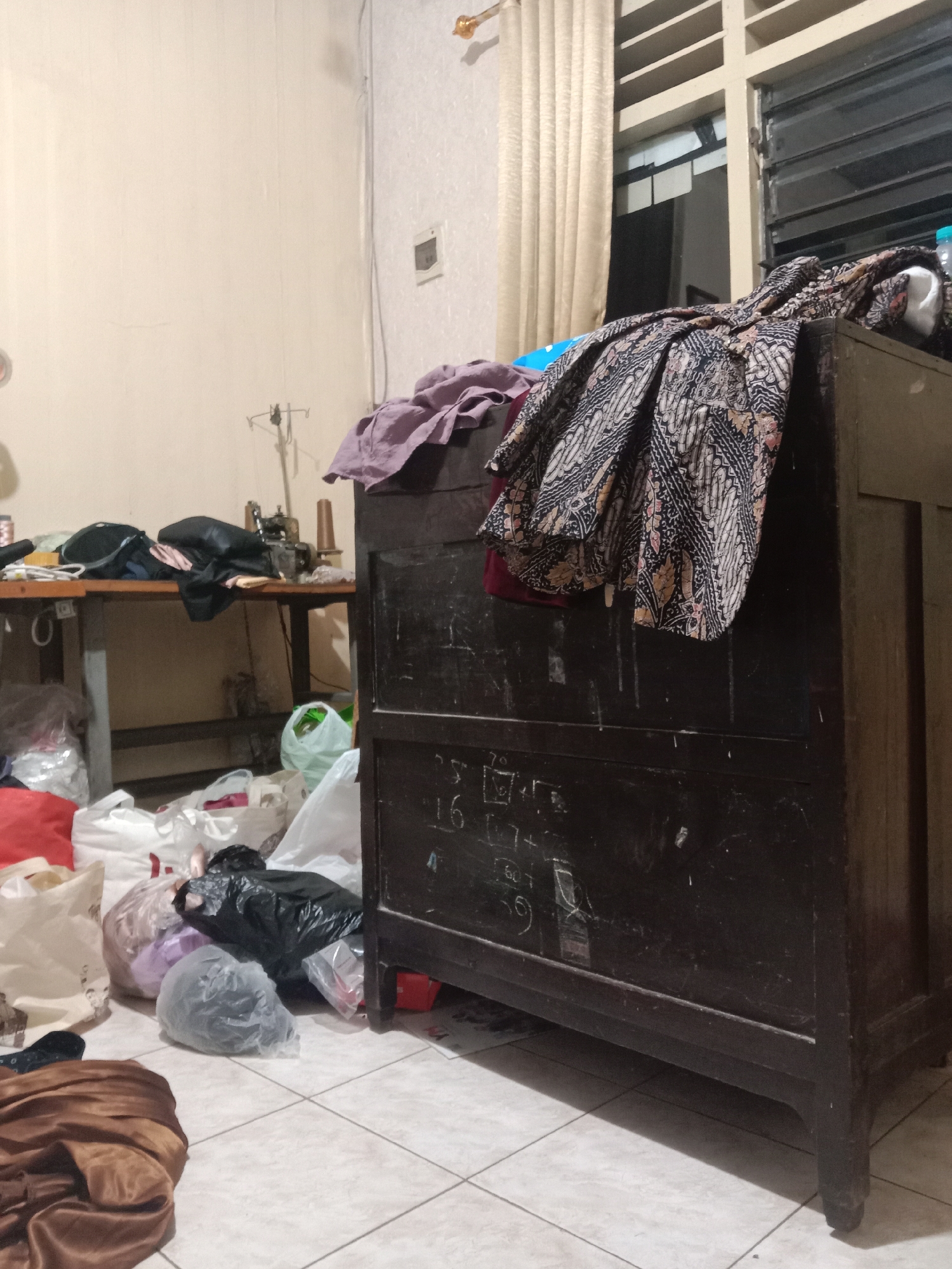 The image depicts a cluttered room, possibly a workshop or storage area, located in Madiun, Indonesia. In the foreground, piles of various bags and fabric are scattered across a tiled floor. A dark wooden chest of drawers occupies the right side of the frame, with a patterned fabric draped over its top. The wall behind is a neutral color. To the left, a sewing machine sits on a table, also surrounded by fabric and bags. A clothes rack with a purple garment is visible near the chest. The room appears to be dimly lit, with a window and curtains visible in the upper right corner. The overall impression is one of disarray and a space dedicated to sewing or fabric work.