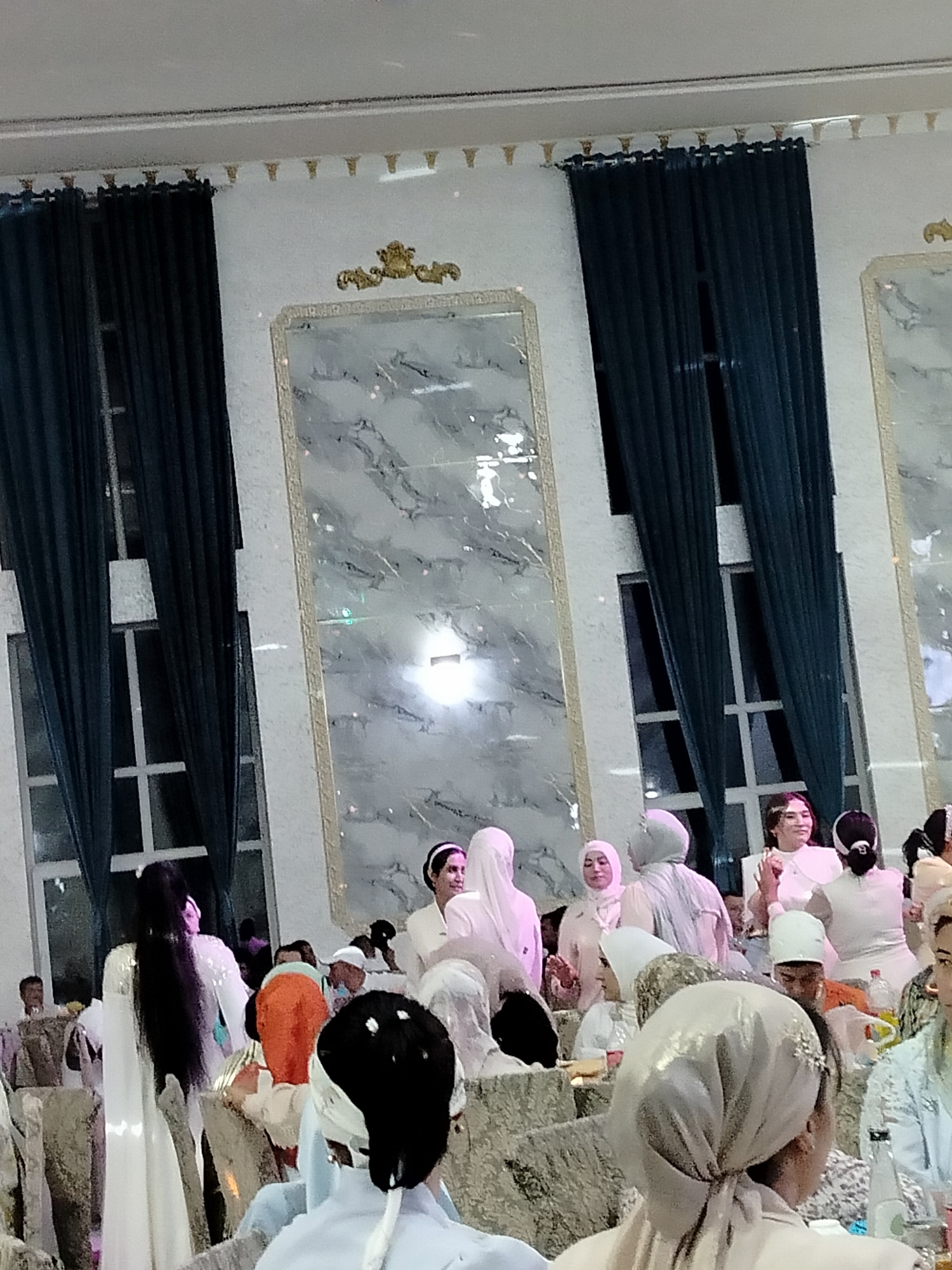 The image depicts an indoor gathering of people, likely a celebration or event, at a venue in Ақсуабат, Kazakhstan. The room features tall, dark blue curtains on either side of large, window-like structures with white frames. The walls are white and adorned with ornate gold trim along the top and a large, decorative gold emblem above a prominent, framed panel with a marbled pattern. This panel is centrally located on the back wall. In the foreground and midground, various individuals are visible, predominantly women, many wearing headscarves and white or light-colored attire. Some people are seated, while others are standing and interacting. The overall atmosphere appears festive.
