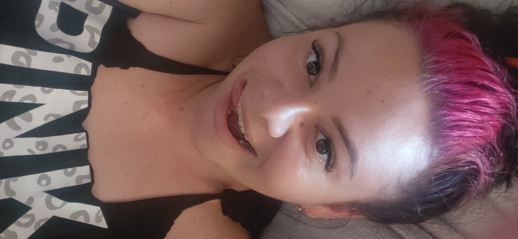 A woman with pink and black hair, wearing a black top with "PINK" written on it in a leopard print font, is lying down and looking at the camera. Her tongue is sticking out and touching her lips. The image is a selfie, taken from a close-up perspective. The background appears to be a bed or couch. There are no discernible landmarks or contextual clues to indicate the location as Marynivka, Ukraine.