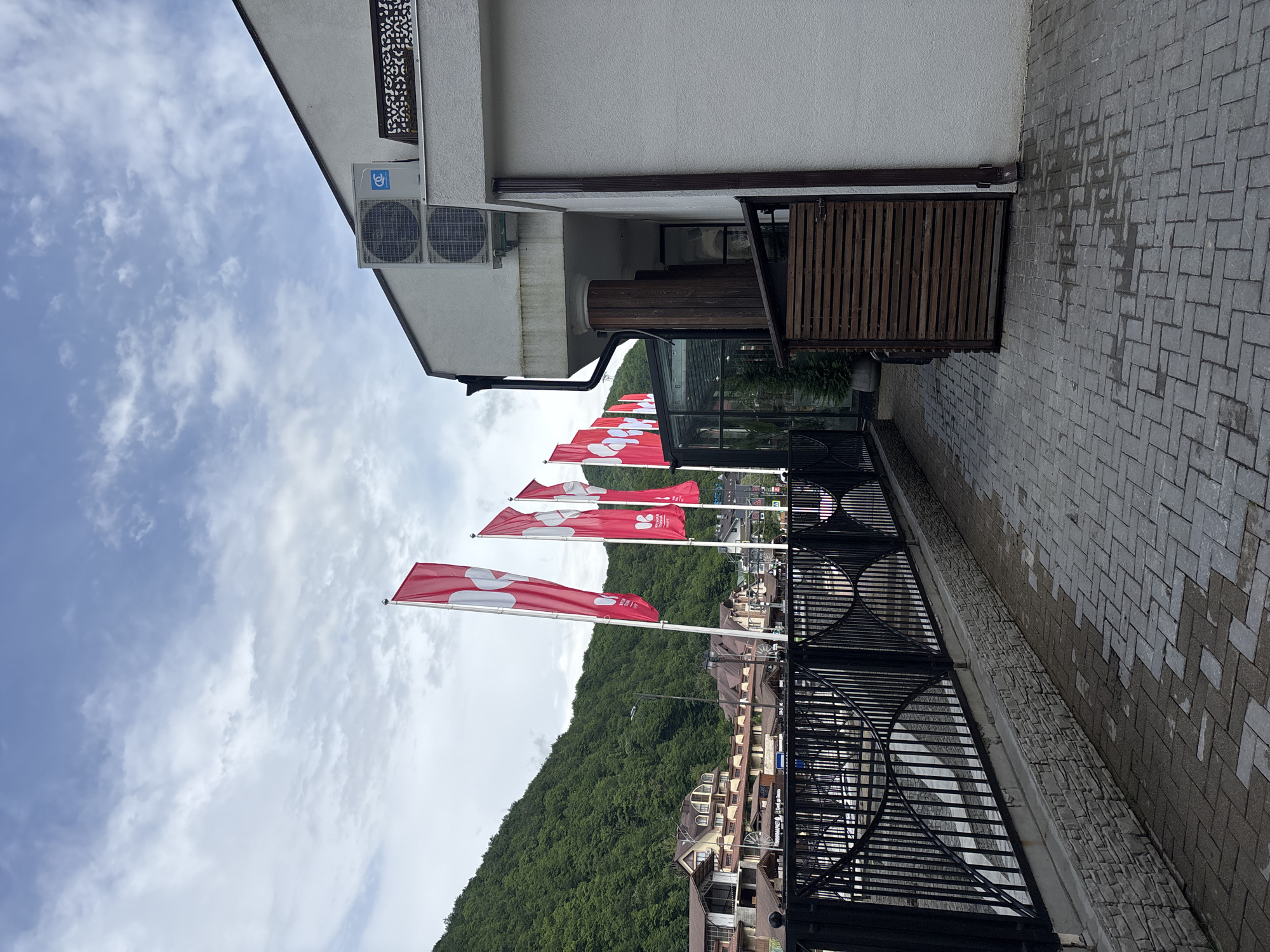The image depicts a street scene in Estosadok, Russia, with a focus on a modern building and a row of red and white flags. The building is white with two air conditioning units mounted on its exterior wall. A black metal fence borders a paved walkway, which shows signs of recent rain with darker patches on the pavers. Beyond the fence, a wooded hillside with buildings nestled within it rises in the background. A series of red flags with white circular logos are displayed prominently on flagpoles, lining the walkway and adding a decorative element to the scene. The sky is cloudy. There are no people visible in the image.