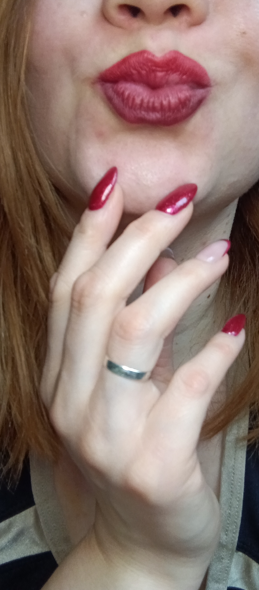 The image is a close-up, chest-up shot of a person, focusing on their mouth and a hand. The person has fair skin and reddish-brown hair, visible on the sides and bottom left. Their lips are prominently featured, pursed into a "kissing" shape and covered in a matte red lipstick. A small portion of the nose is visible just above the upper lip.
A hand is raised towards the face, with four fingers visible. The thumb, index, and middle fingers have long, pointed nails painted in a glossy, shimmering dark red polish. The ring finger has a French manicure style nail with a light pink base and a dark red tip, also shaped similarly. A simple, smooth silver band ring is worn on the ring finger. The skin on the hand and face shows natural texture, including some visible pores and minor blemishes.
A small section of clothing is visible on the lower right, featuring black and light-colored stripes, possibly part of a shirt or top. A plain, light-colored strap is also seen near the neck/shoulder area on the right. The overall impression is a playful or affectionate pose. There is no visible text in the image.