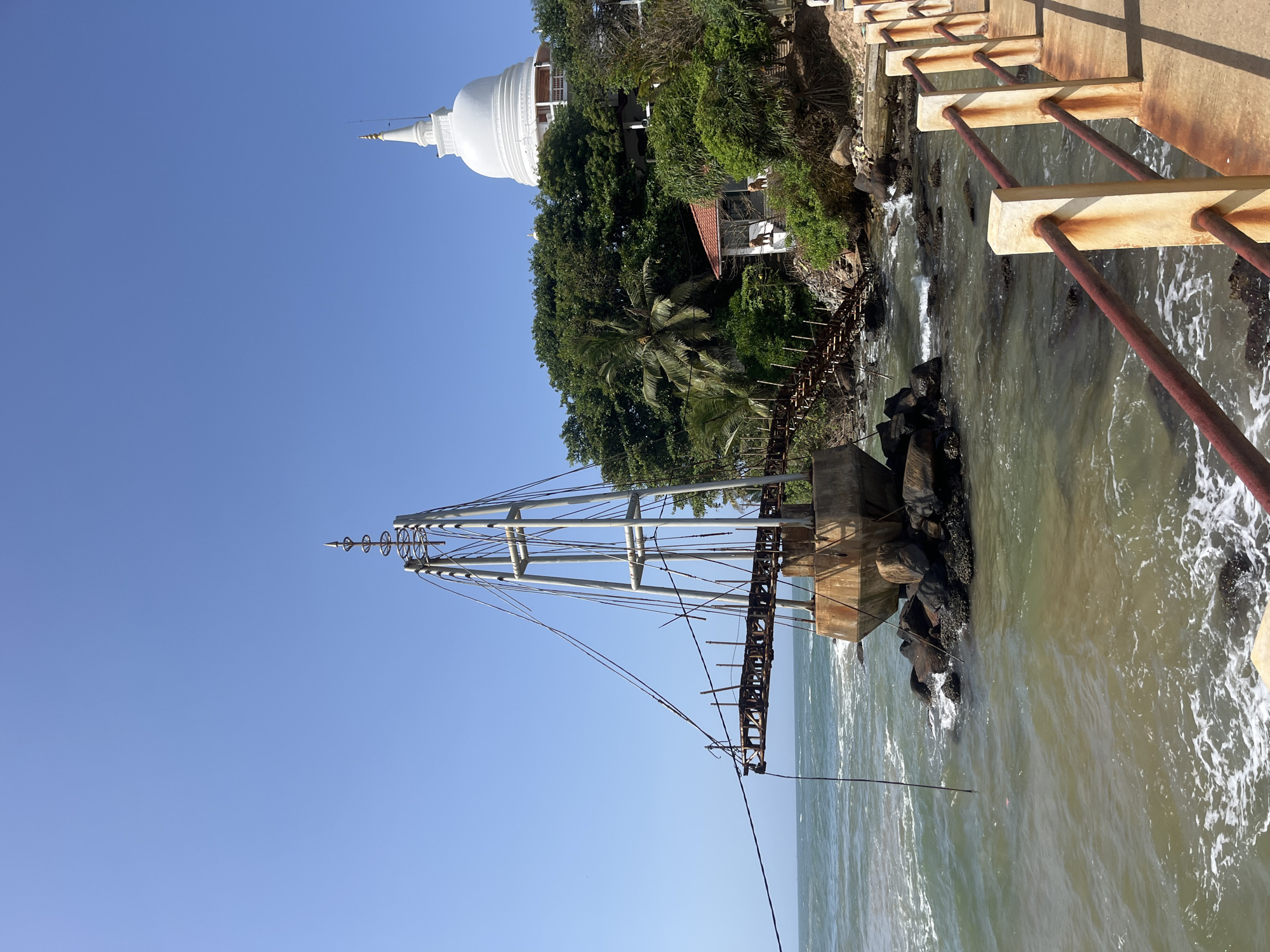 This image depicts the suspended pedestrian bridge at the Weherahena Buddhist Temple in Matara, Sri Lanka. The bridge, constructed from metal and concrete, extends over the ocean towards a rocky shore. It is anchored to a large, concrete foundation situated among dark, weathered rocks in the water. The bridge itself appears to be in a state of disrepair, with visible rust and missing structural elements, suggesting it is no longer in active use. A tall, skeletal metal tower rises from the concrete foundation, with numerous cables extending outwards, once supporting the bridge.

In the background, lush green trees and foliage cover a hillside. Perched atop this hill is the prominent white stupa of the Weherahena Buddhist Temple, its gleaming dome a stark contrast to the natural surroundings. The clear blue sky serves as the backdrop for the entire scene. The foreground features a concrete walkway with a rusted metal railing, indicating a designated viewing area or path along the coastline. The ocean displays gentle waves, with some white foam lapping against the rocks and the base of the bridge structure. The lighting suggests it is daytime, with a bright sun casting shadows. No people are visible in the image.