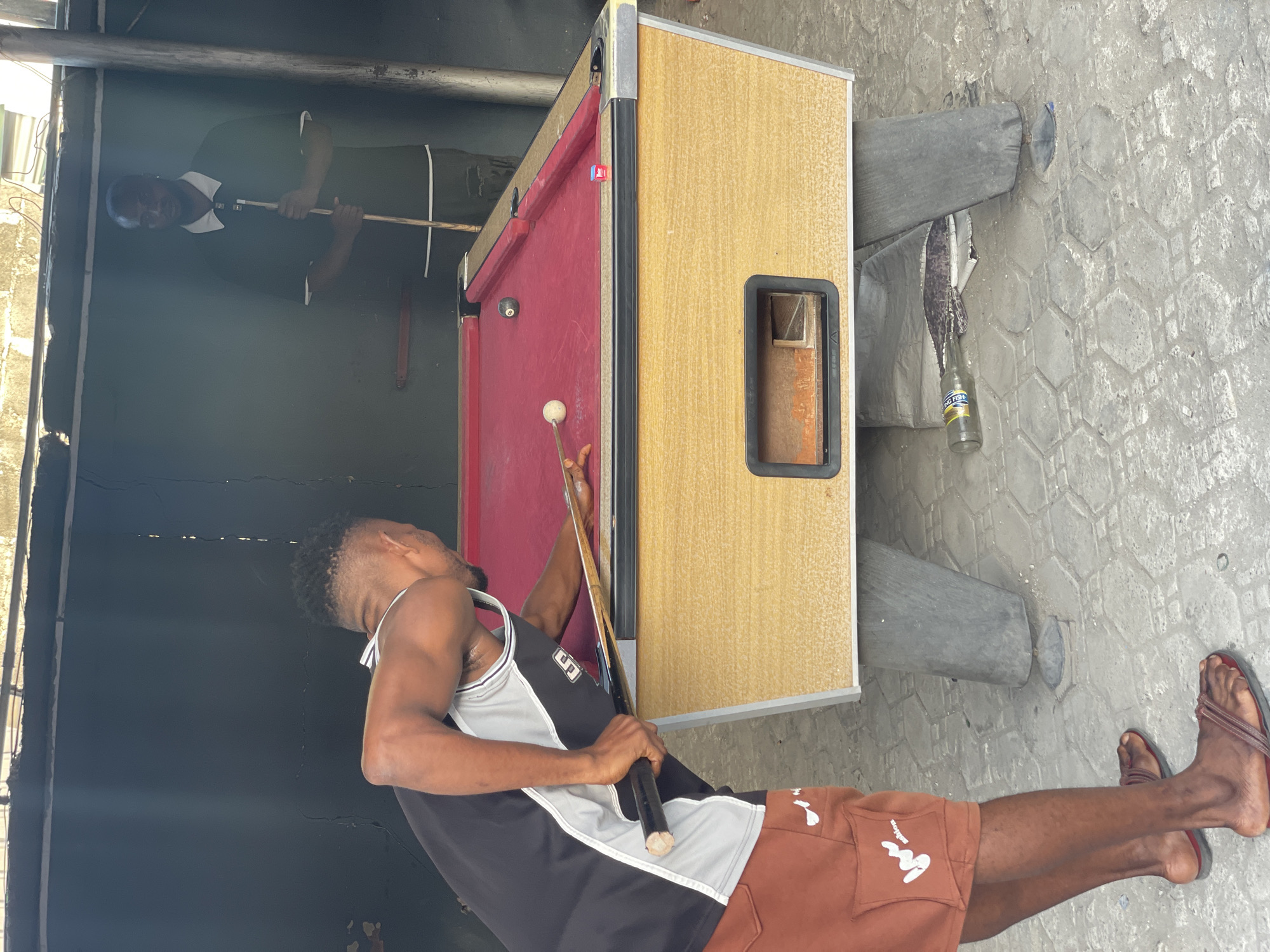 The image depicts an outdoor scene, likely in a casual recreational area in Ado, Nigeria, featuring a pool table.
A man in a grey and black tank top and brown shorts with white decorative patches is aiming a pool cue at a white cue ball on the red felt of the table. Another person in a dark shirt with a white collar is visible in the background, standing near the pool table holding a cue as well.
The pool table itself is tan colored with a red felt surface. The table is supported by cylindrical metal legs. A brown beer bottle sits near one of the legs on the grey hexagonal tiled ground. The background includes a dark wall.