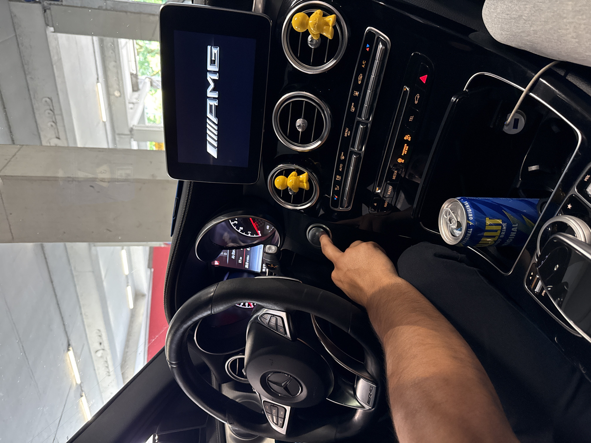 The image shows the interior of a Mercedes-AMG car. The infotainment screen displays the AMG logo. The dashboard features air vents, two of which have yellow, abstract figures attached. A man's hand is pressing the start button. A can of "Original Energy Drink" is in a cup holder. The steering wheel has the Mercedes logo in the center. The car is likely in a garage, given the background with concrete pillars. It is impossible to ascertain whether the image was taken in Gelsenkirchen, Germany with the provided information.